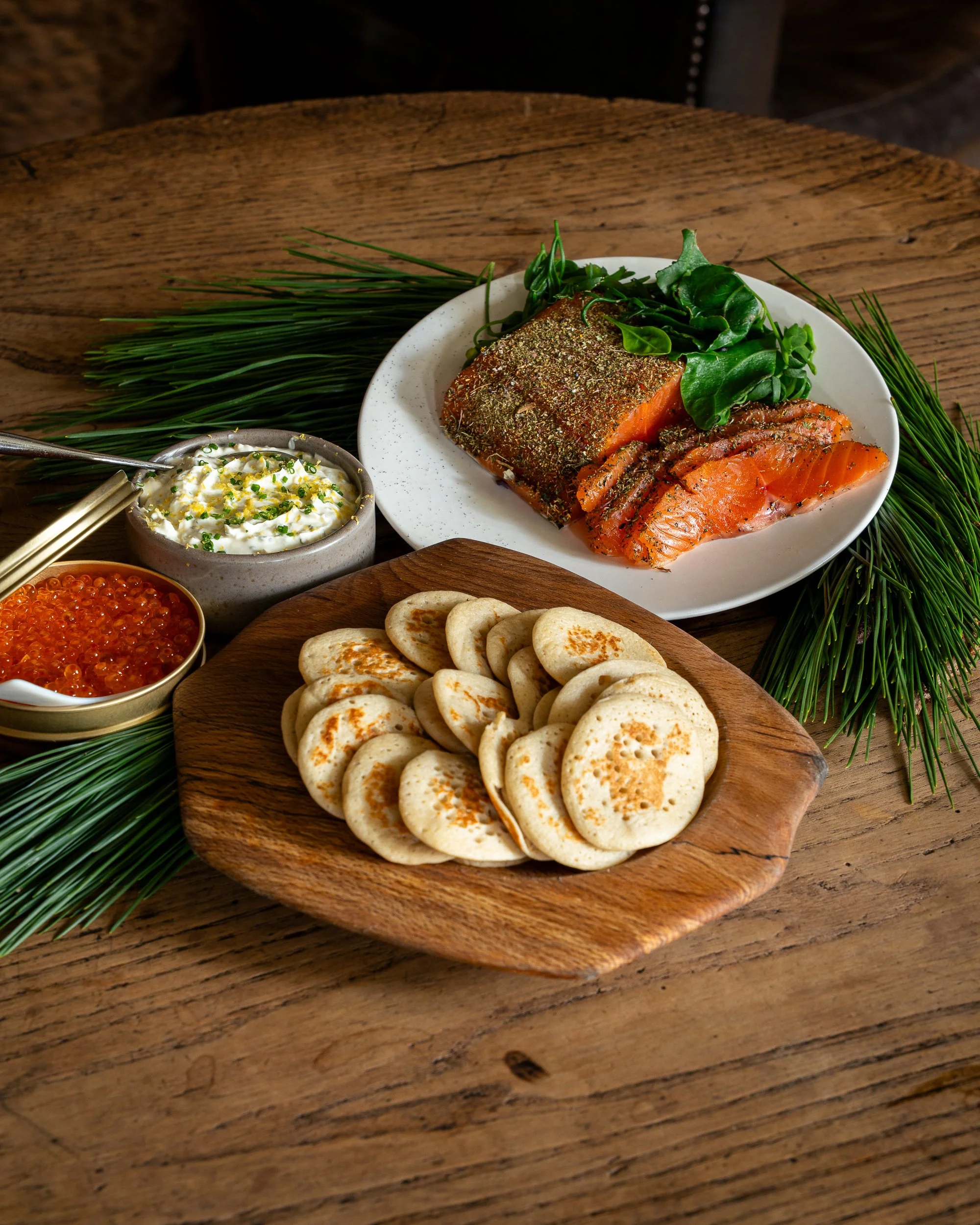 Close-up of a wooden table with a white plate holding smoked salmon, leafy greens, and herbs, alongside bowls of caviar, cream cheese, and a wooden platter with small toasted bread rounds, garnished with fresh pine branches.