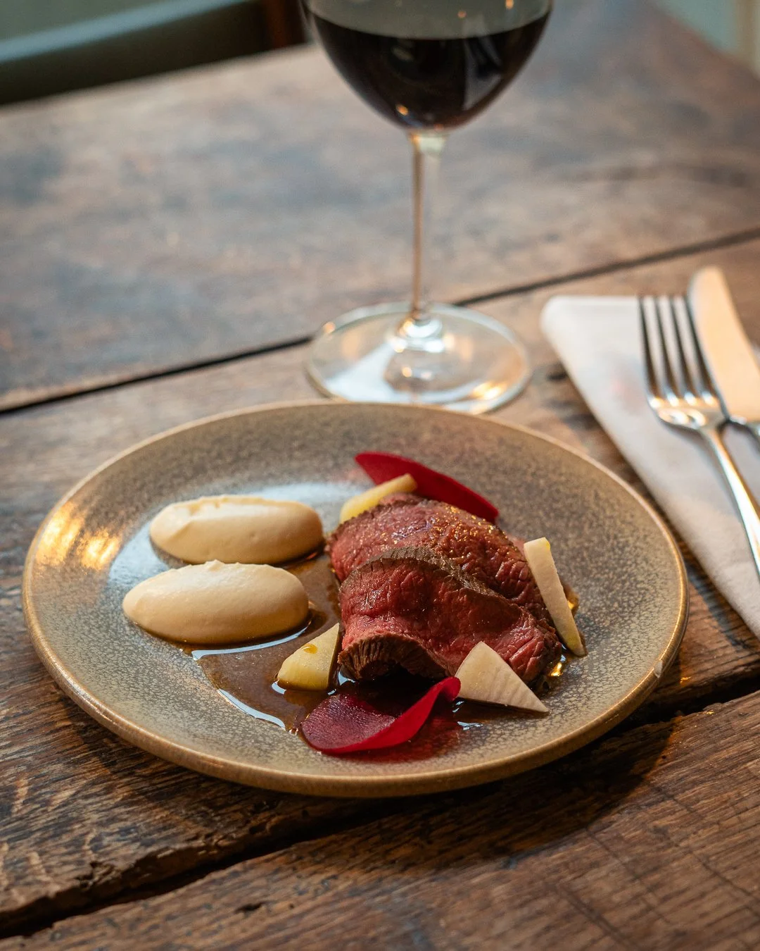 A plate of cooked venison with sauce, turnip puree, and garnishes, next to a glass of red wine on a wooden table.