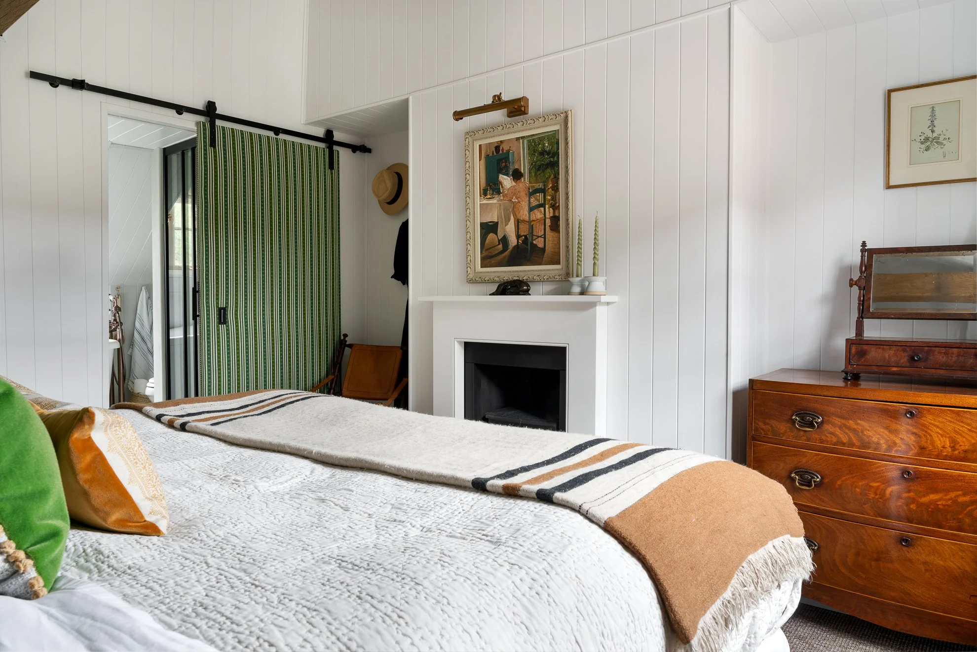 A cozy bedroom with white paneled walls, a bed with a white quilt and colorful pillows, a vintage wooden dresser, a white fireplace mantel with decorative candles, framed artwork, and a green-striped curtain covering a doorway.