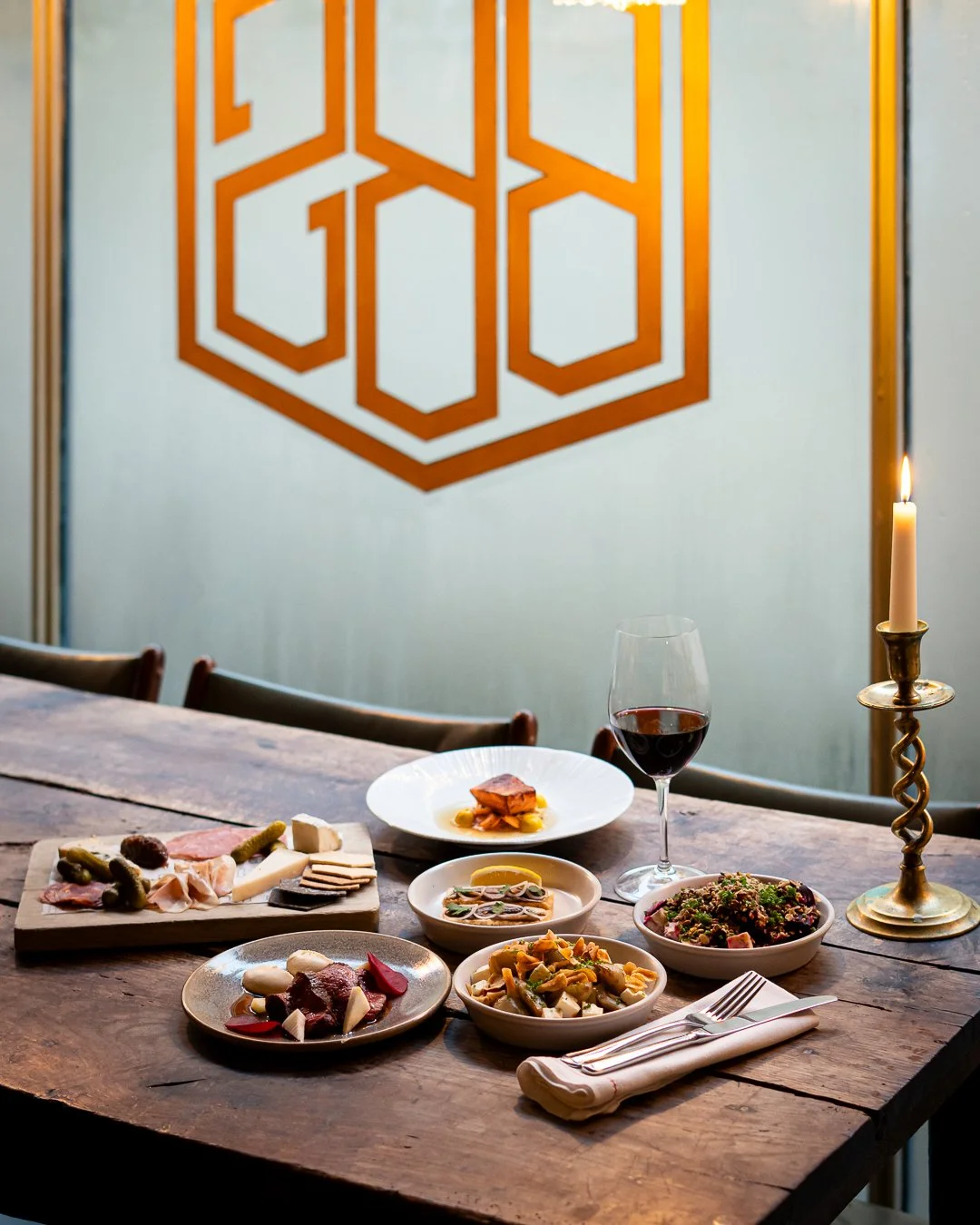 A rustic wooden dining table set with various dishes, a glass of red wine, and a lit candlestick. A white plate with grilled vegetables, a bowl of pasta, and other small dishes are arranged on the table. The background features a wall with a geometri