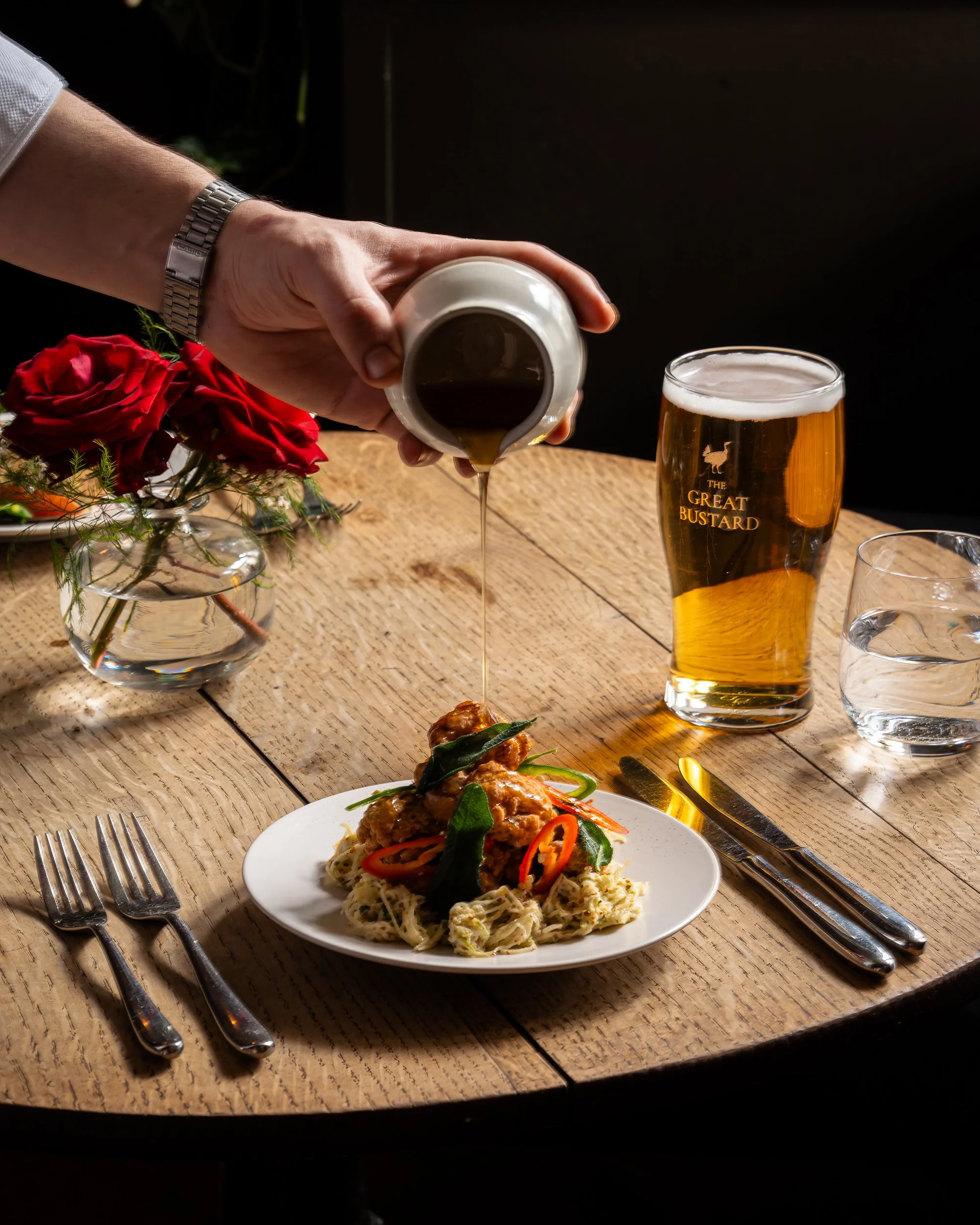 A person pouring brown sauce over a plate of noodles and vegetables, with a glass of beer, water, and a floral arrangement on a wooden table.