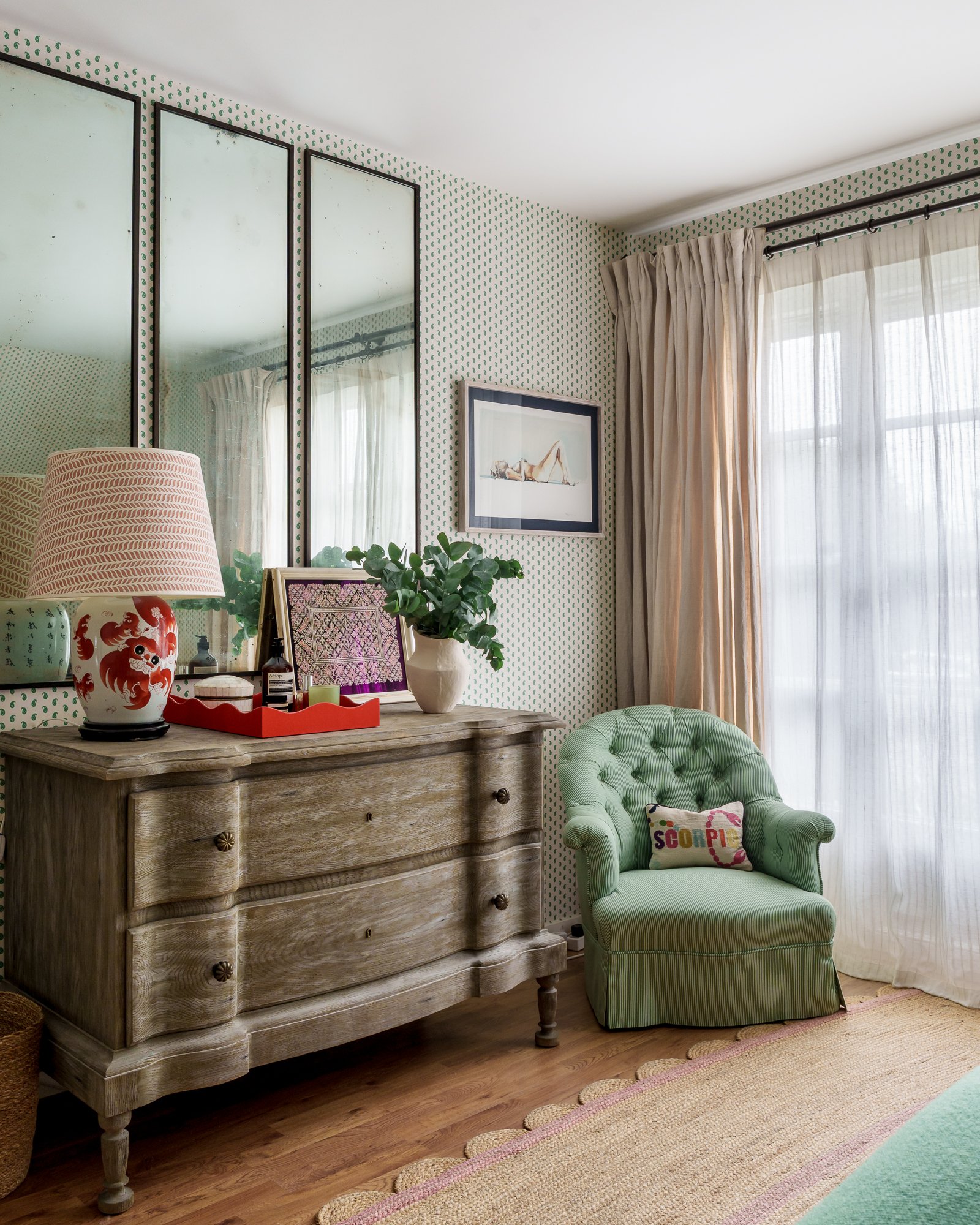 A room corner with a wooden dresser, a green upholstered armchair with a decorative pillow, a table lamp, reflective mirrors, a framed art of a reclining woman, and curtains covering a window.