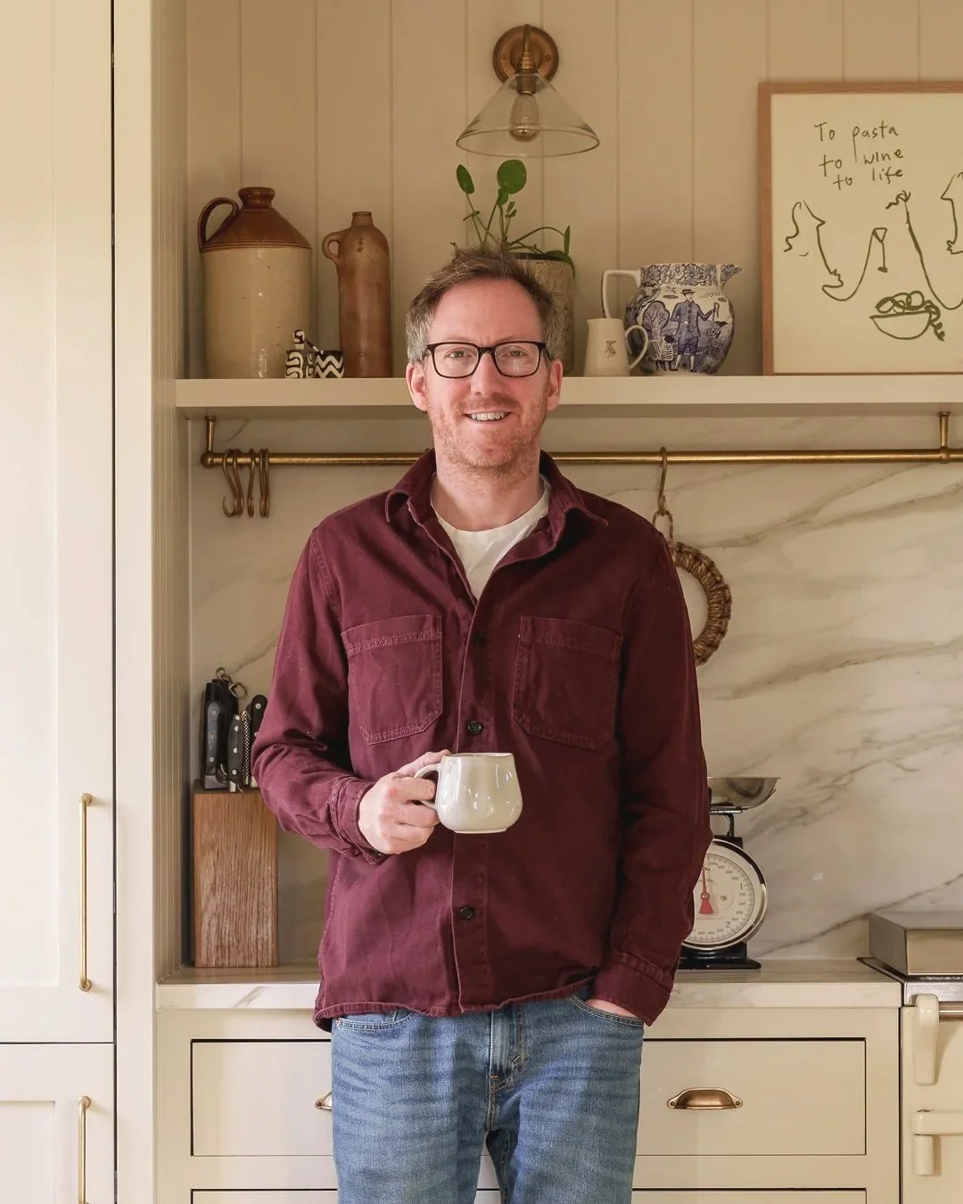 A man with glasses and a beard holding a mug in a kitchen with beige cabinets and decorative items on open shelves.