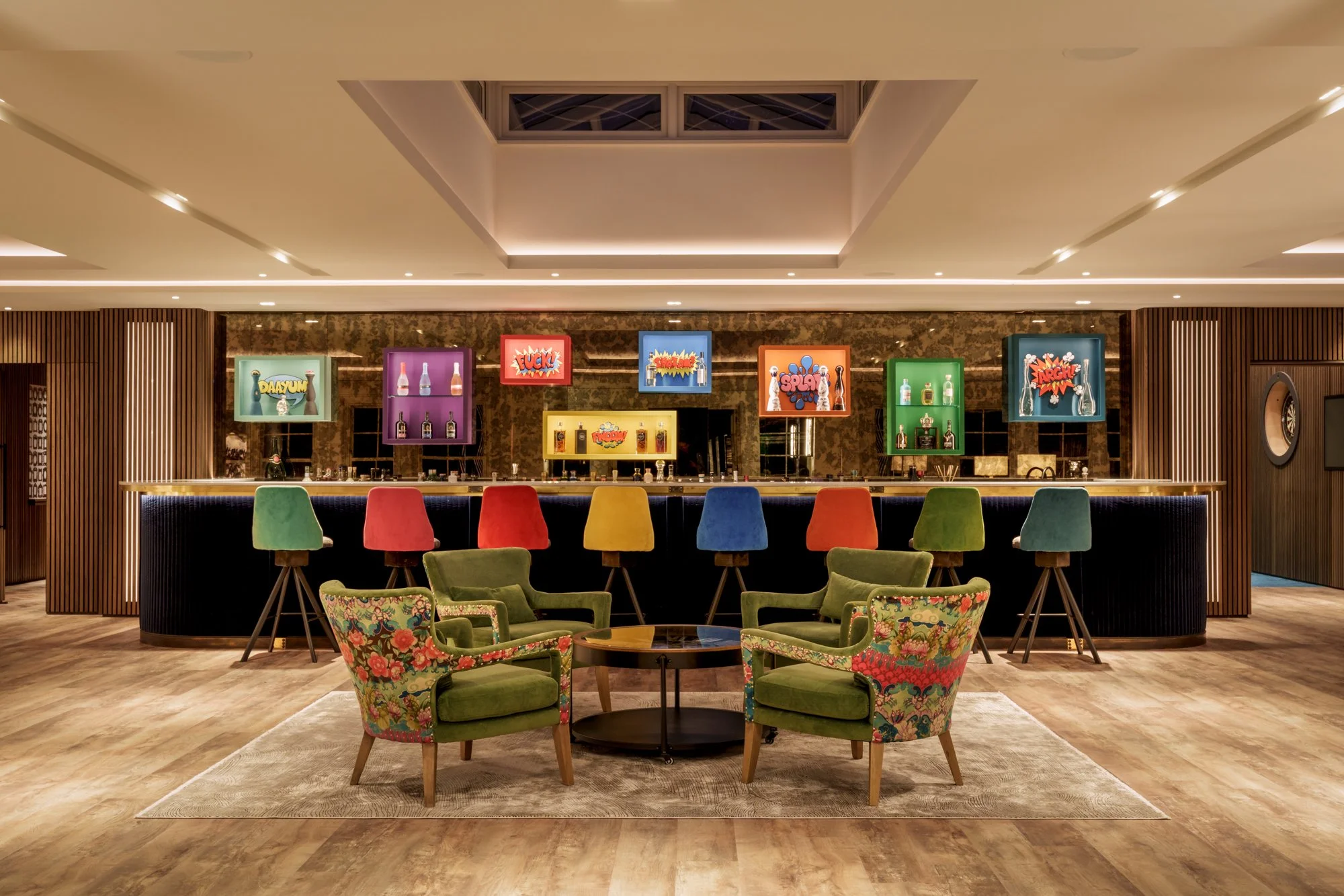 Interior of a bar with a curved counter, colorful chairs, vintage armchairs with floral pattern around a small round table, and illuminated shelves with bottles and comic-style art.
