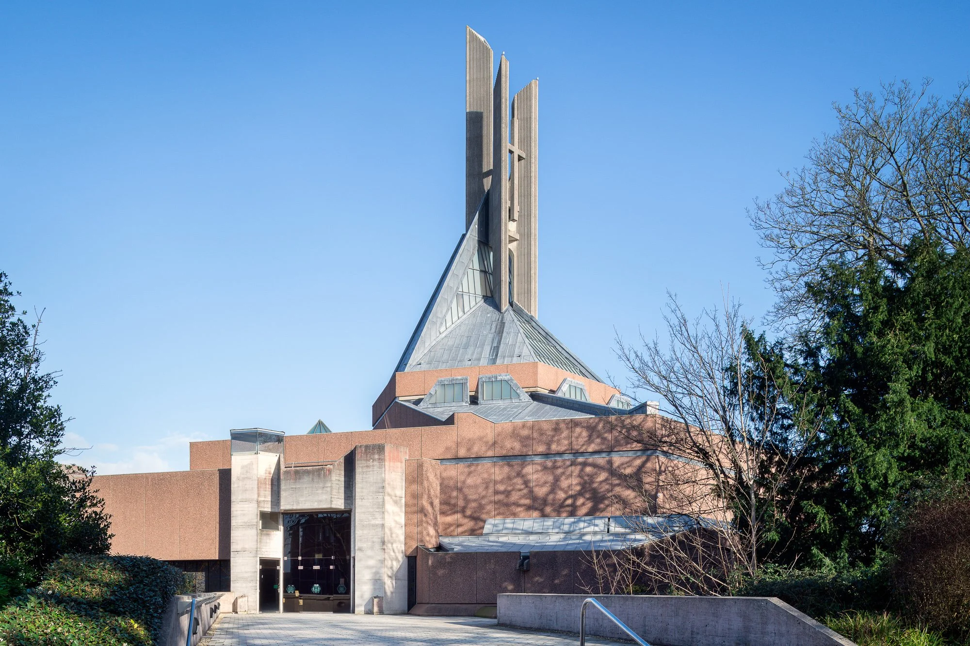 Modern church with a tall, pointed, wooden steeple against a clear blue sky, surrounded by trees and low-rise brick and concrete building.