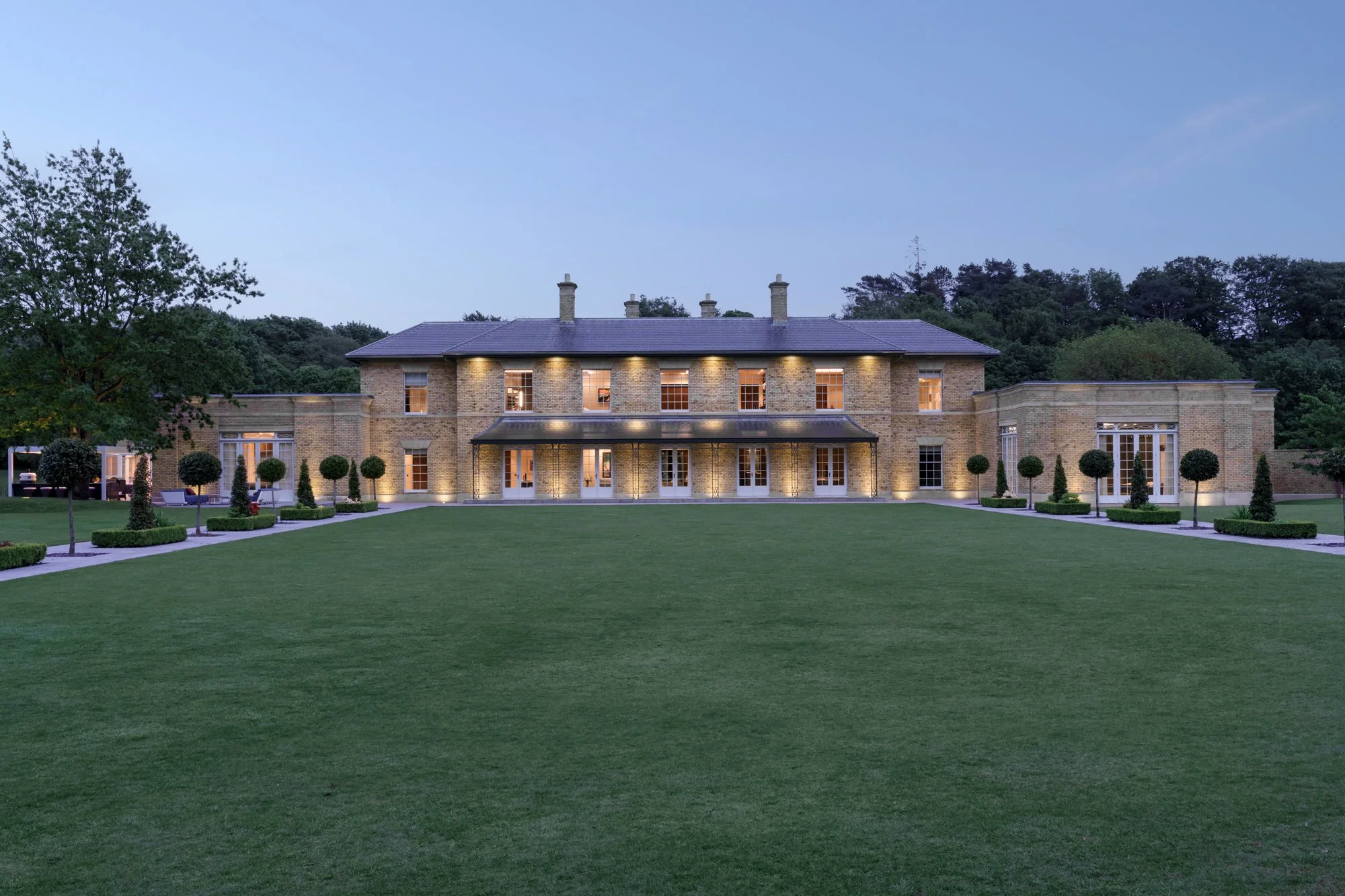 Large brick house with multiple chimneys, illuminated windows, and a spacious well-maintained lawn with trees and shrubs, evening scene with twilight sky.