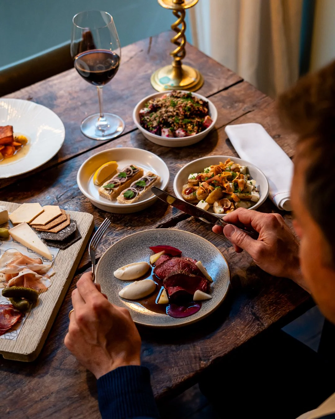 Close-up of a person serving themselves food from a dinner table with various dishes, including sliced meat with sauce, salads, cheese platter, and a glass of red wine.
