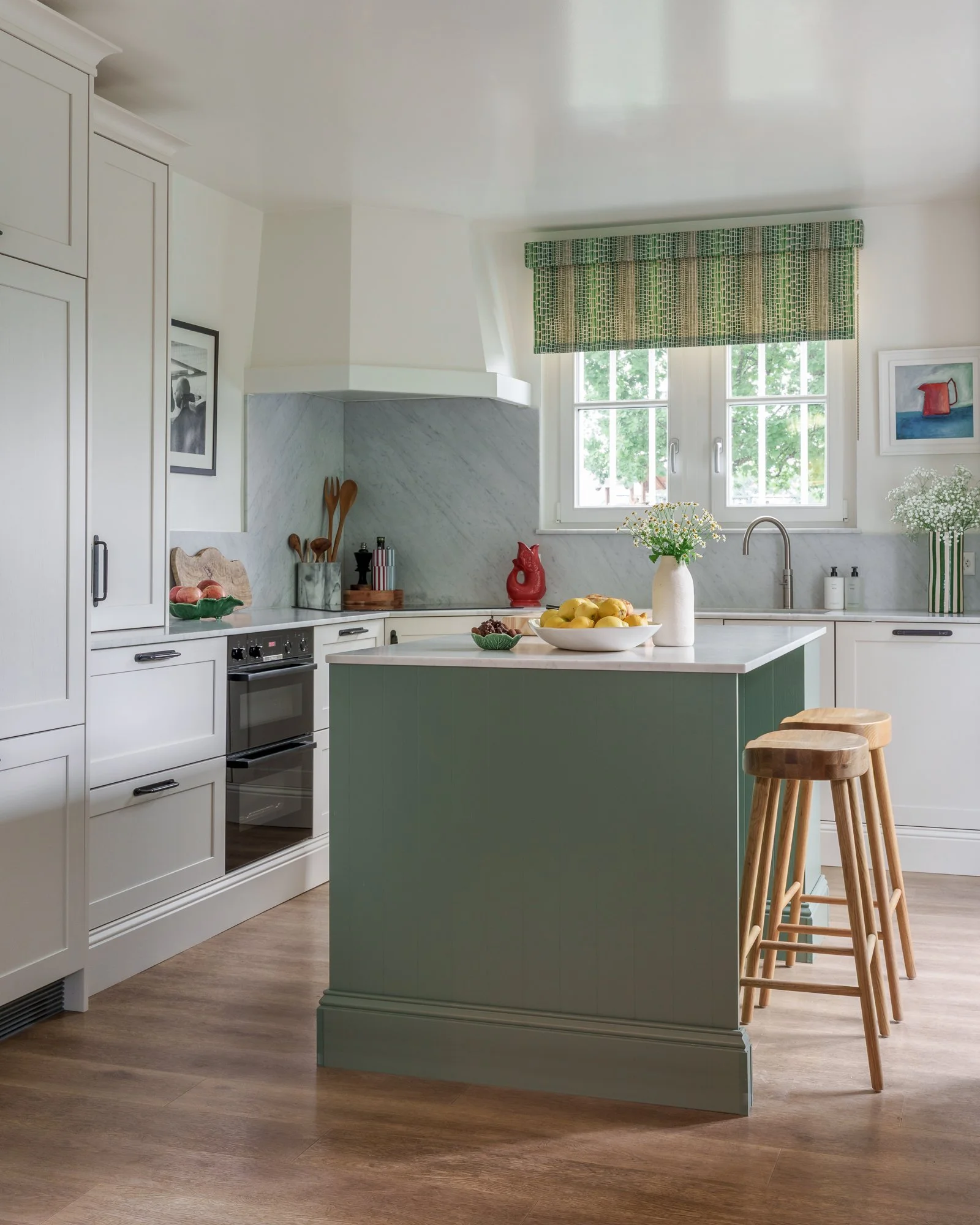 A modern kitchen with white cabinets, wooden stools, green island, marble backsplash, and window with green patterned curtain. Countertop has a bowl of apples, a vase with flowers, and kitchen utensils.