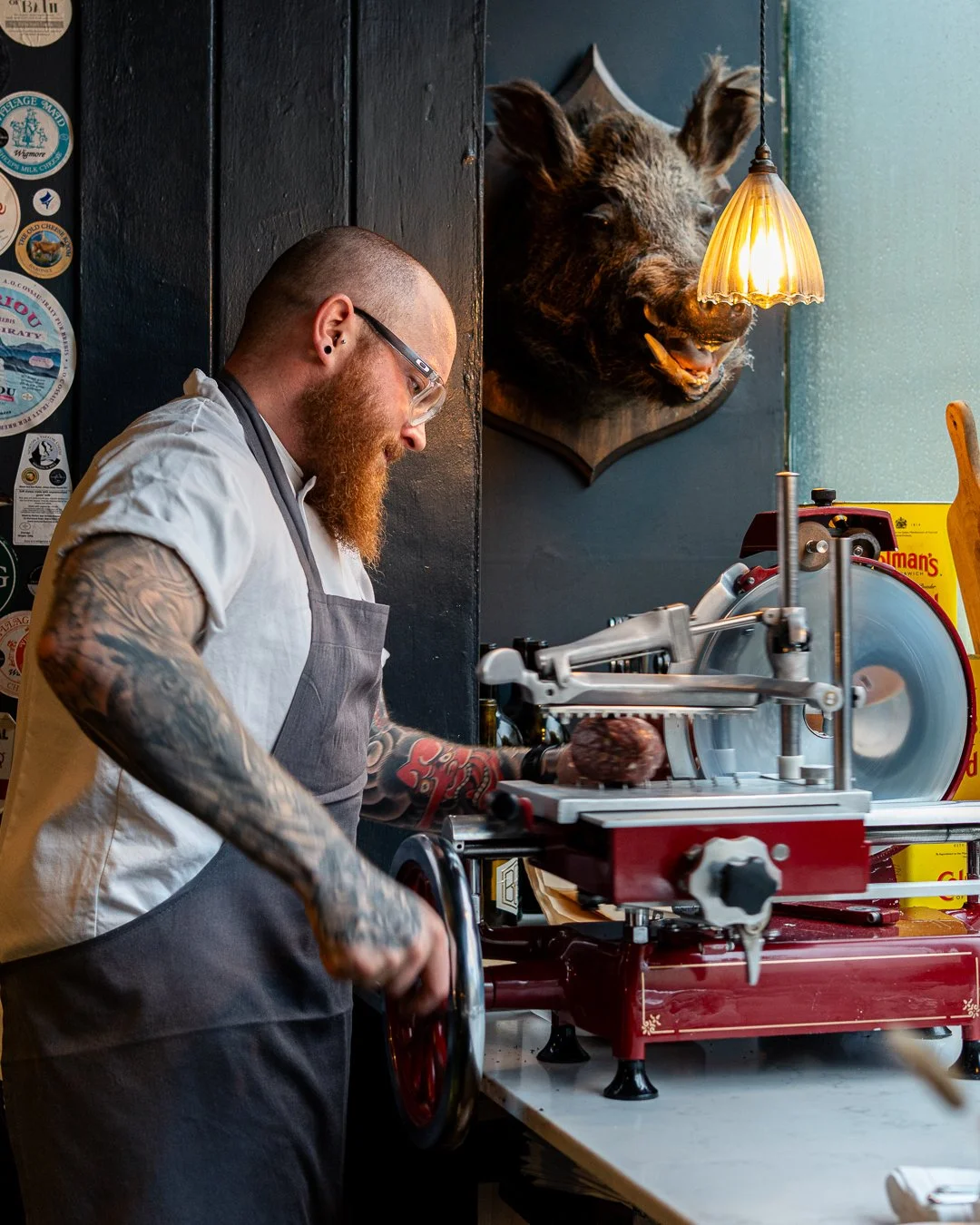 A tattooed chef slices meat on a vintage meat slicer in a rustic kitchen, with a mounted boar head on the wall and decorative stickers on the wall.