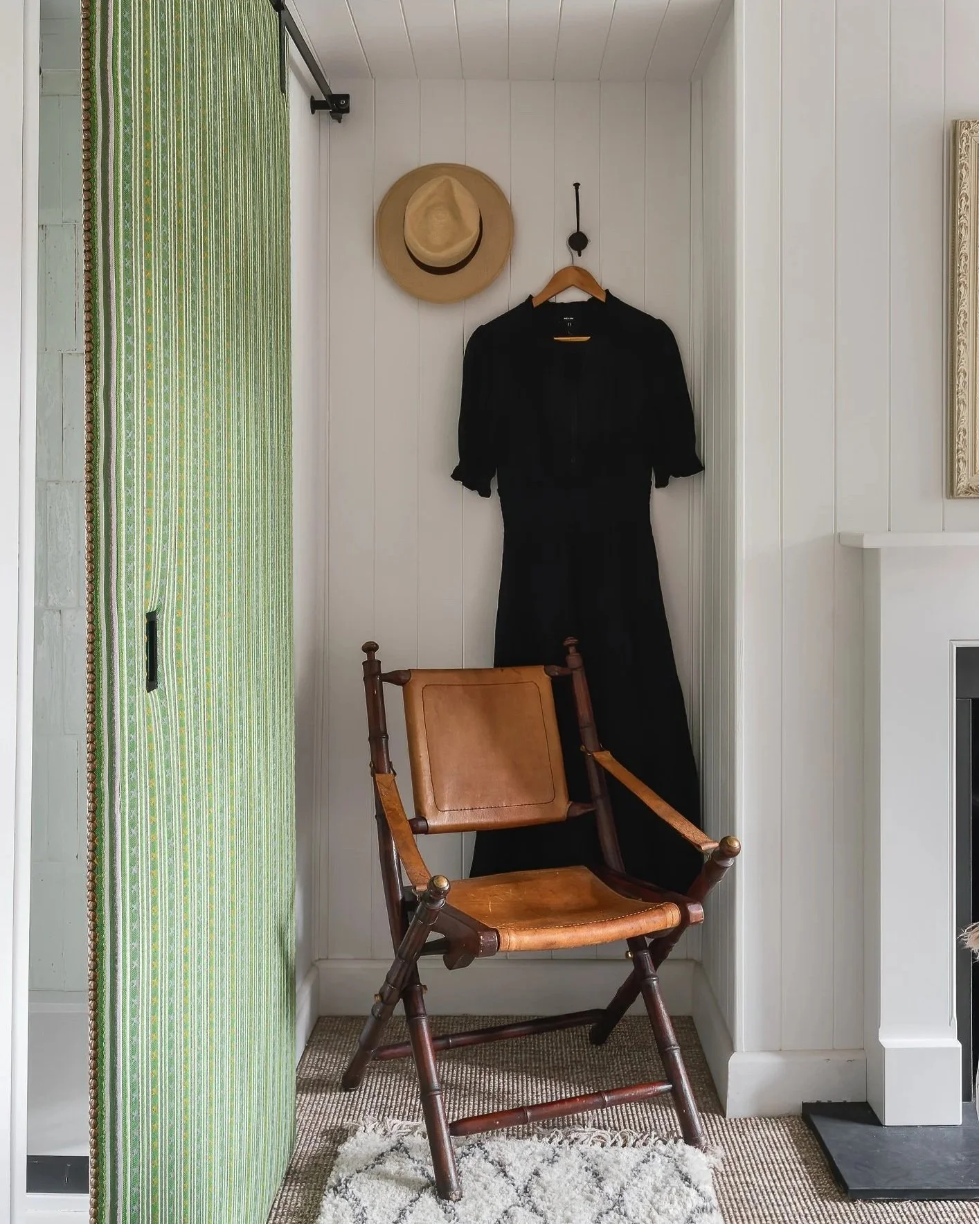 A corner of a room with white paneled walls. A wooden chair with a leather seat is positioned at an angle. Hanging on the wall are a straw hat and a black jacket on a wooden hanger. To the left, part of a sliding door with green striped fabric is visible. The floor is covered in a small brown and beige rug with a white shaggy rug in front of the chair. A part of a fireplace and a black heat vent are also visible.