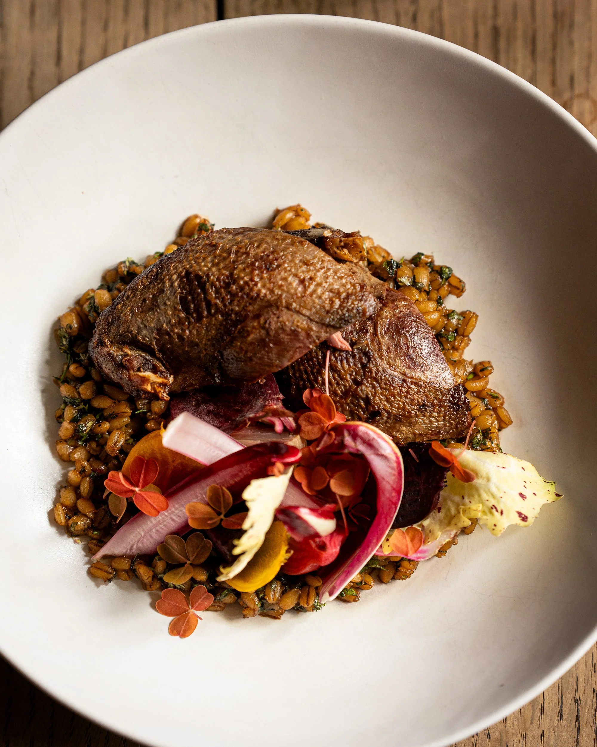 A white bowl contains cooked fish served on a bed of lentils, garnished with edible flowers and thinly sliced vegetables.