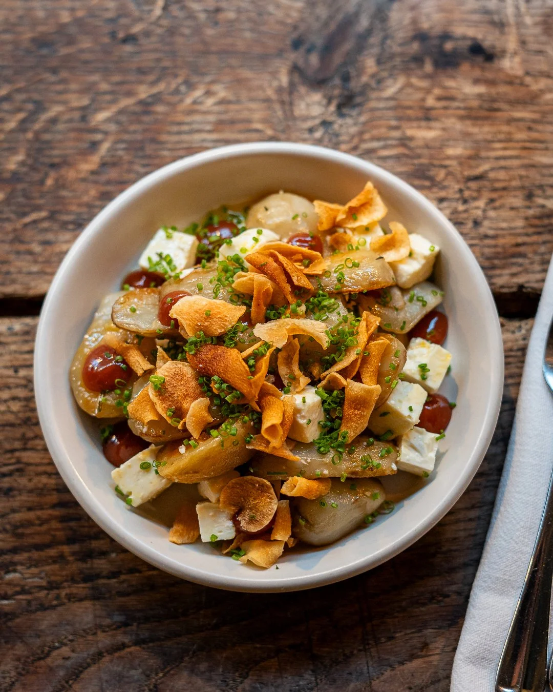 A bowl of artichoke salad topped with fried onion chips, chives, and a smoky sauce on a wooden table.