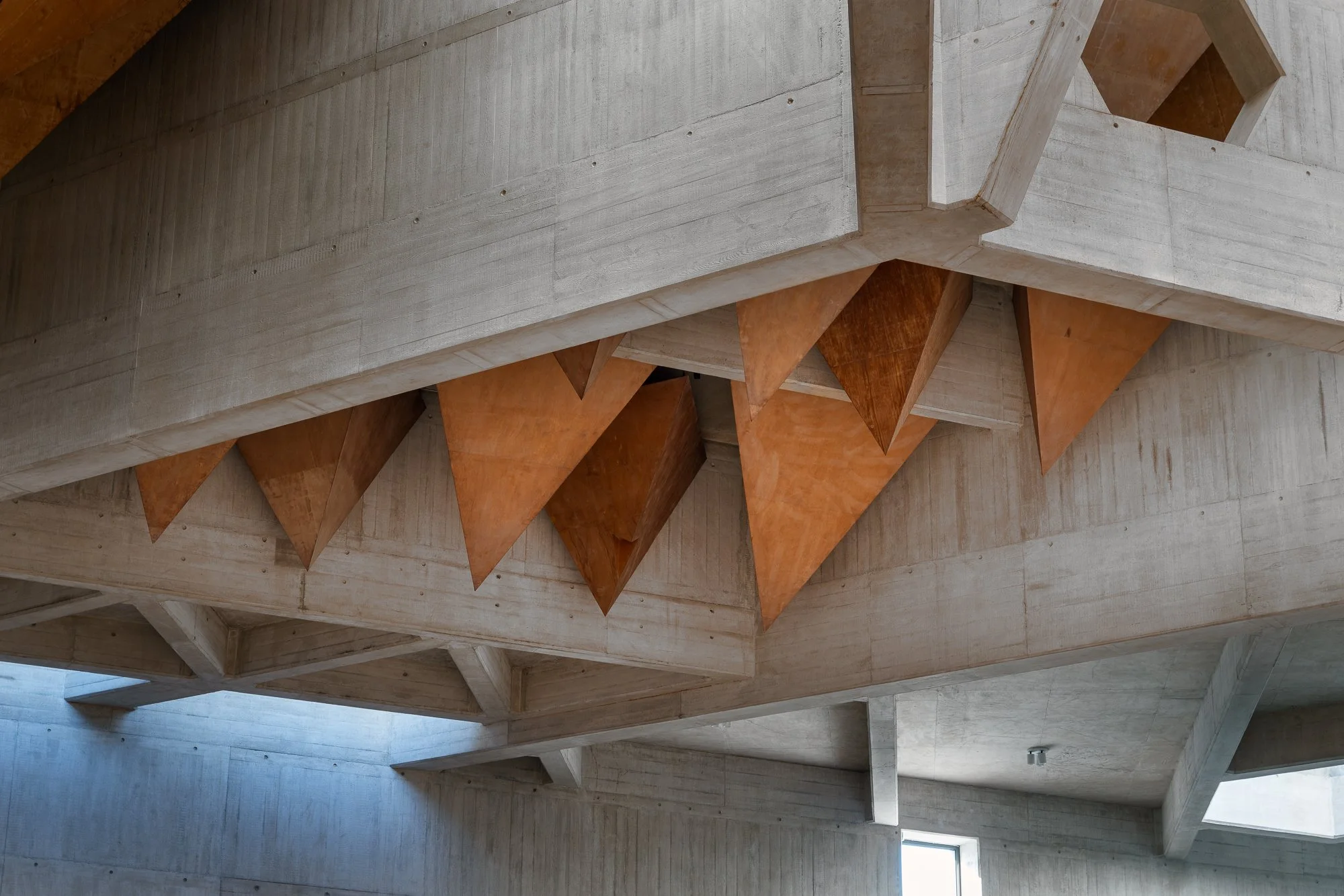 Interior view of a modern building showing concrete walls, wooden beams, and an angular staircase with wooden steps.