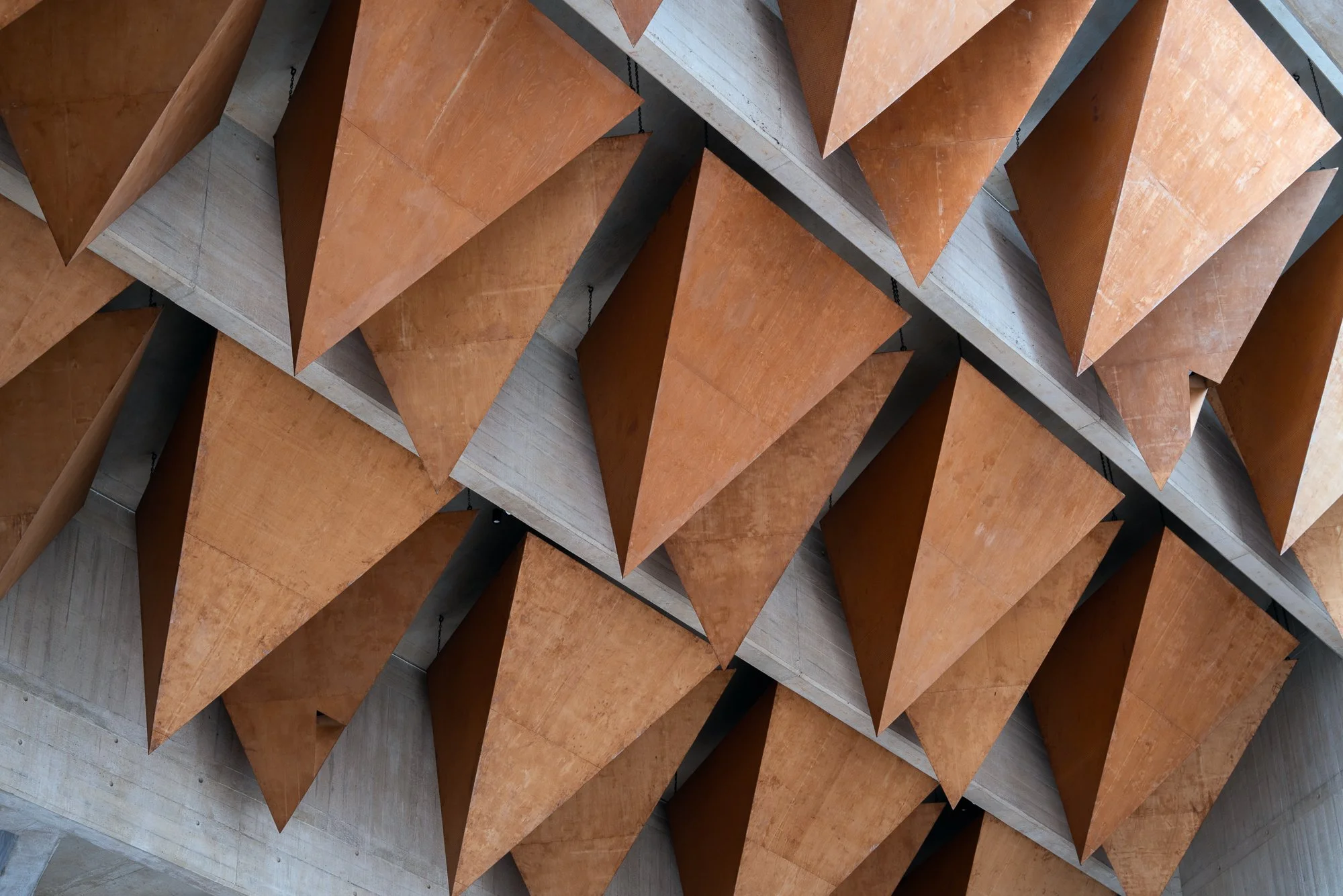 Close-up of a modern architectural wall with angular, rust-colored metal panels arranged in a geometric pattern, supported by visible metal chains and concrete sections.