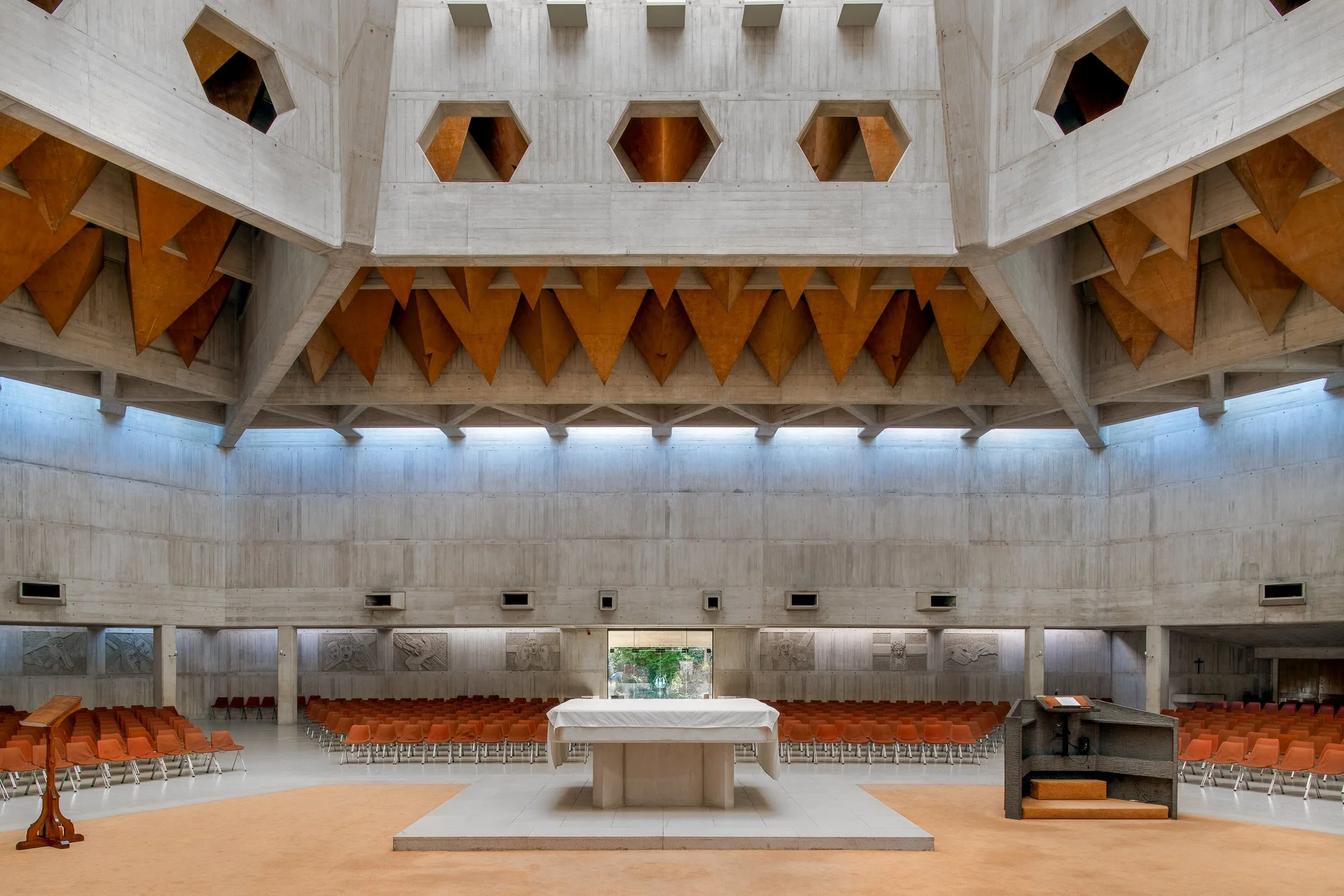 Interior of a modern, concrete building with a high ceiling, featuring geometric wooden accents and rows of orange chairs facing an altar-like area with a table.