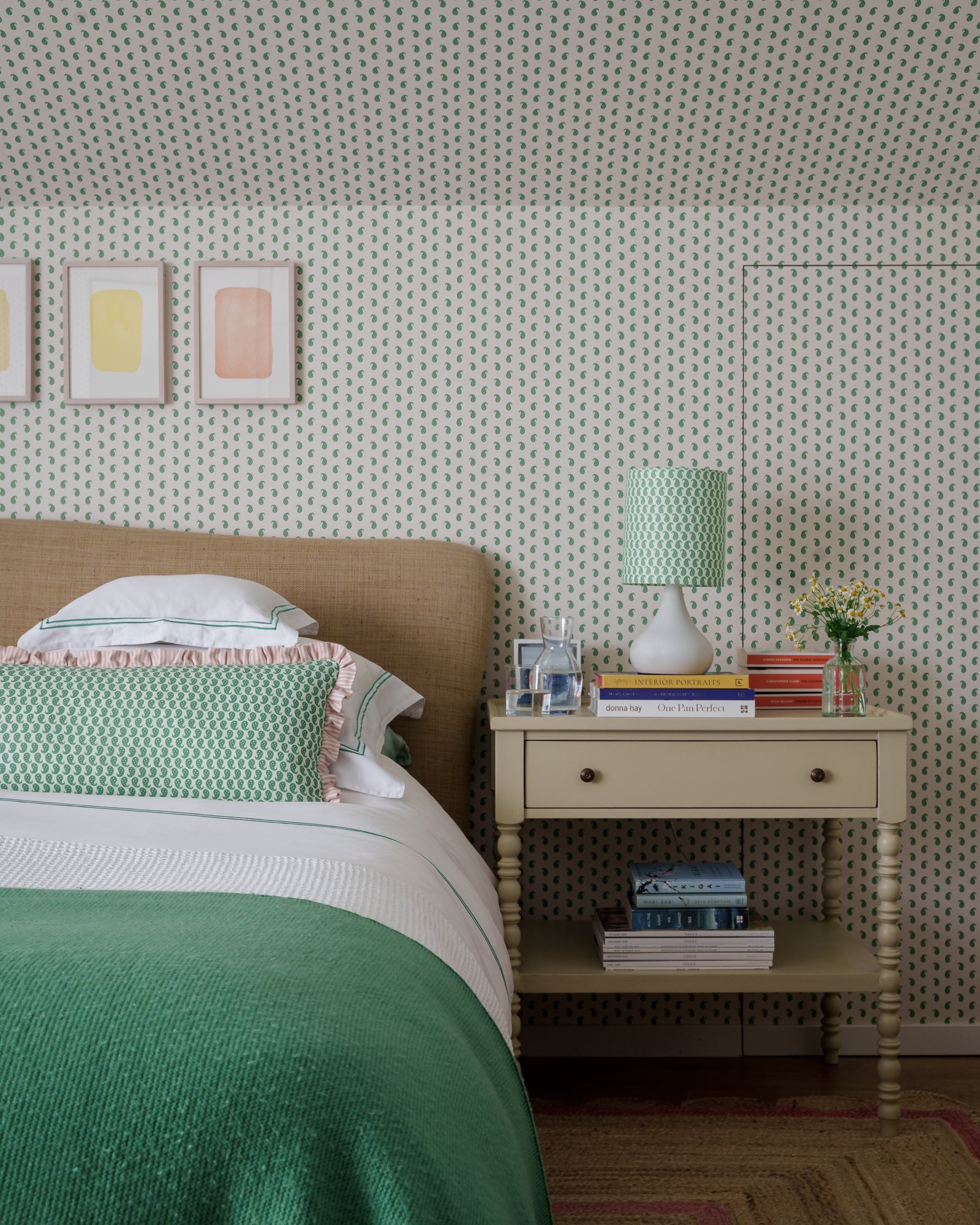 A bedroom with a bed, patterned green and white bedding, a white bedside table with books, a lamp, a glass of water, and a vase with flowers. The wall behind has green patterned wallpaper and framed artwork.