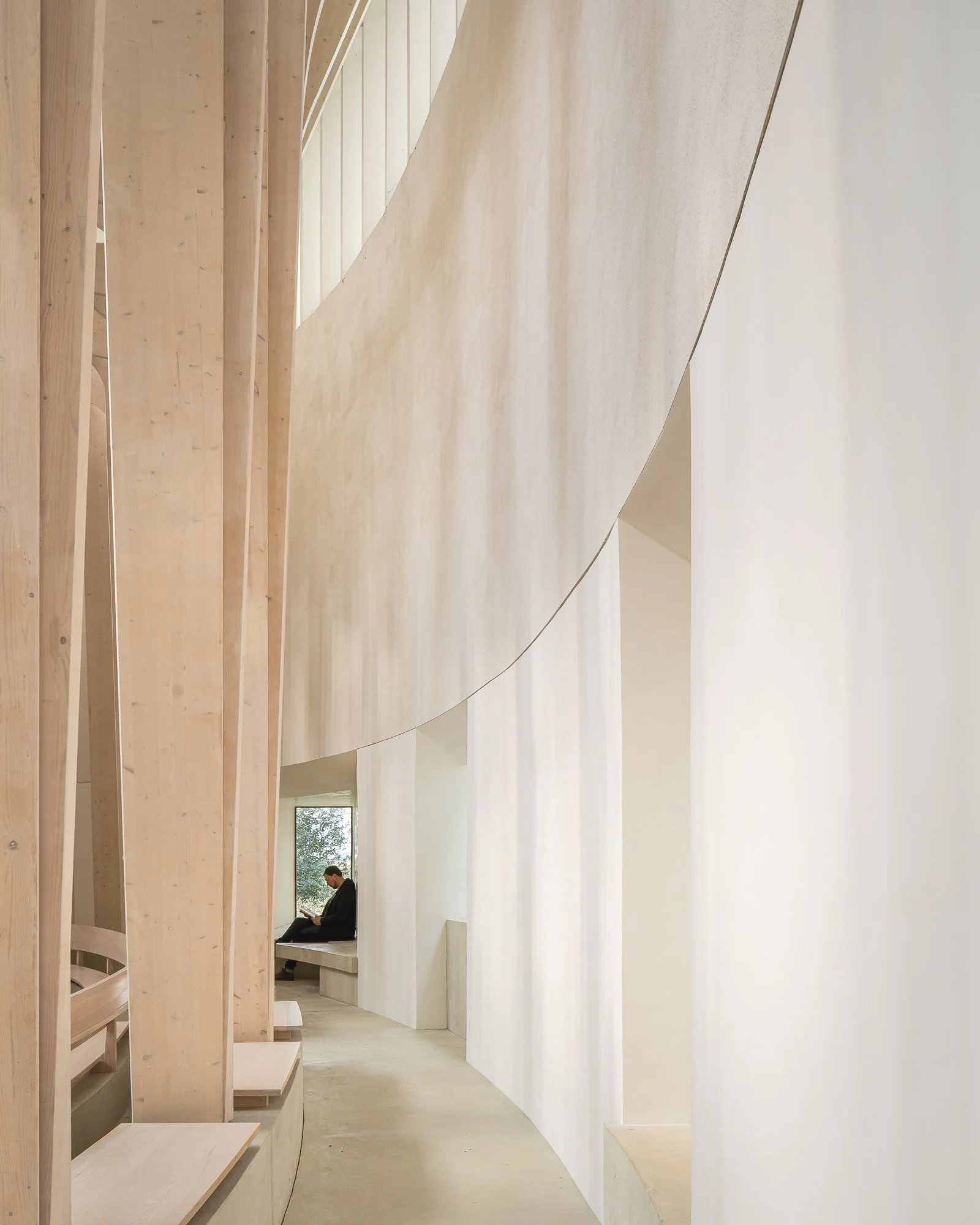Interior view of a modern minimalist space with curved white walls and wooden architectural elements, featuring a person sitting on a bench reading a book near a window.
