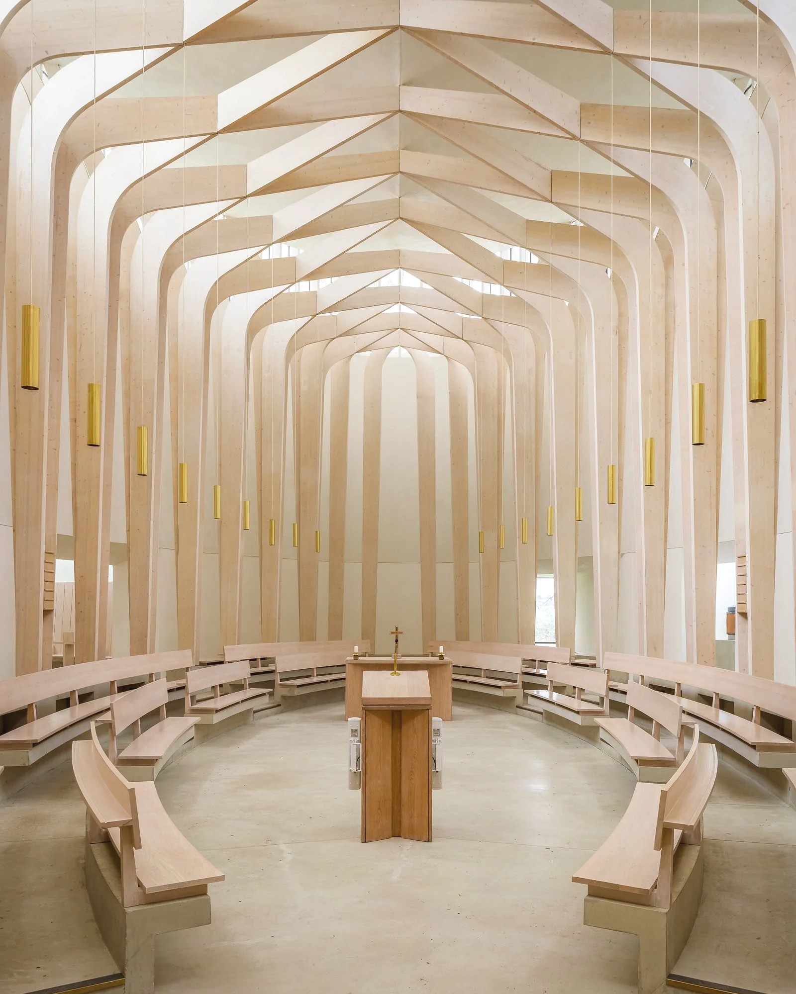 The interior of a church or chapel with a circular seating arrangement and a wooden altar at the center. The space features high, arched wooden ceilings and natural light streaming in from windows behind the altar.