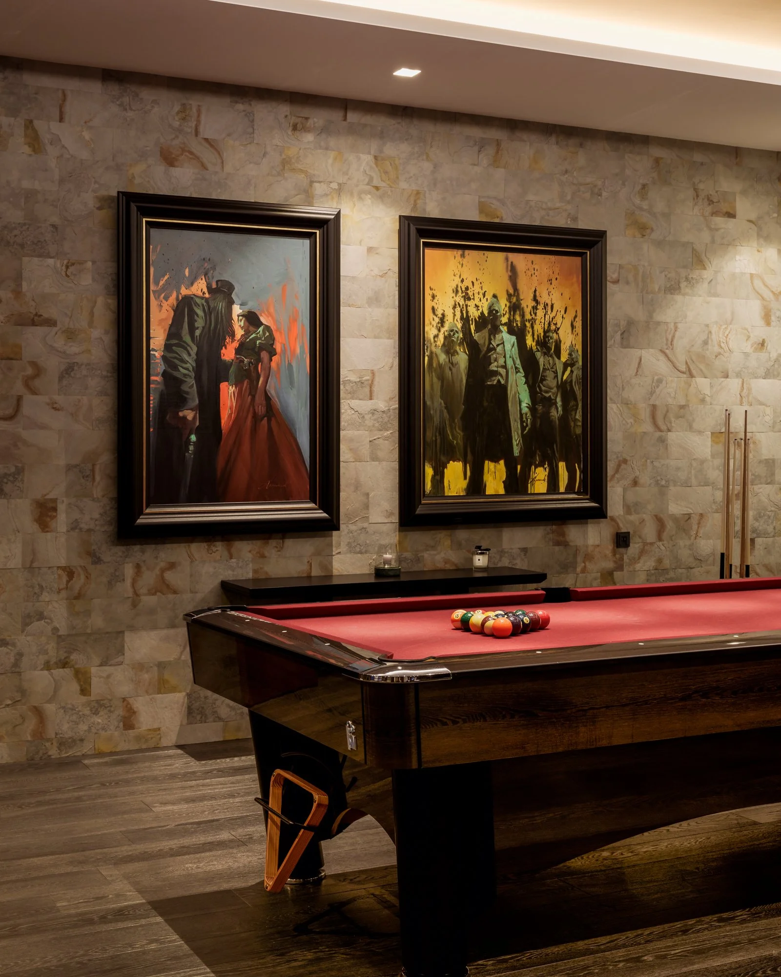 A pool table with billiard balls arranged at the end, a chair next to it, and two framed paintings on a stone wall in the background.