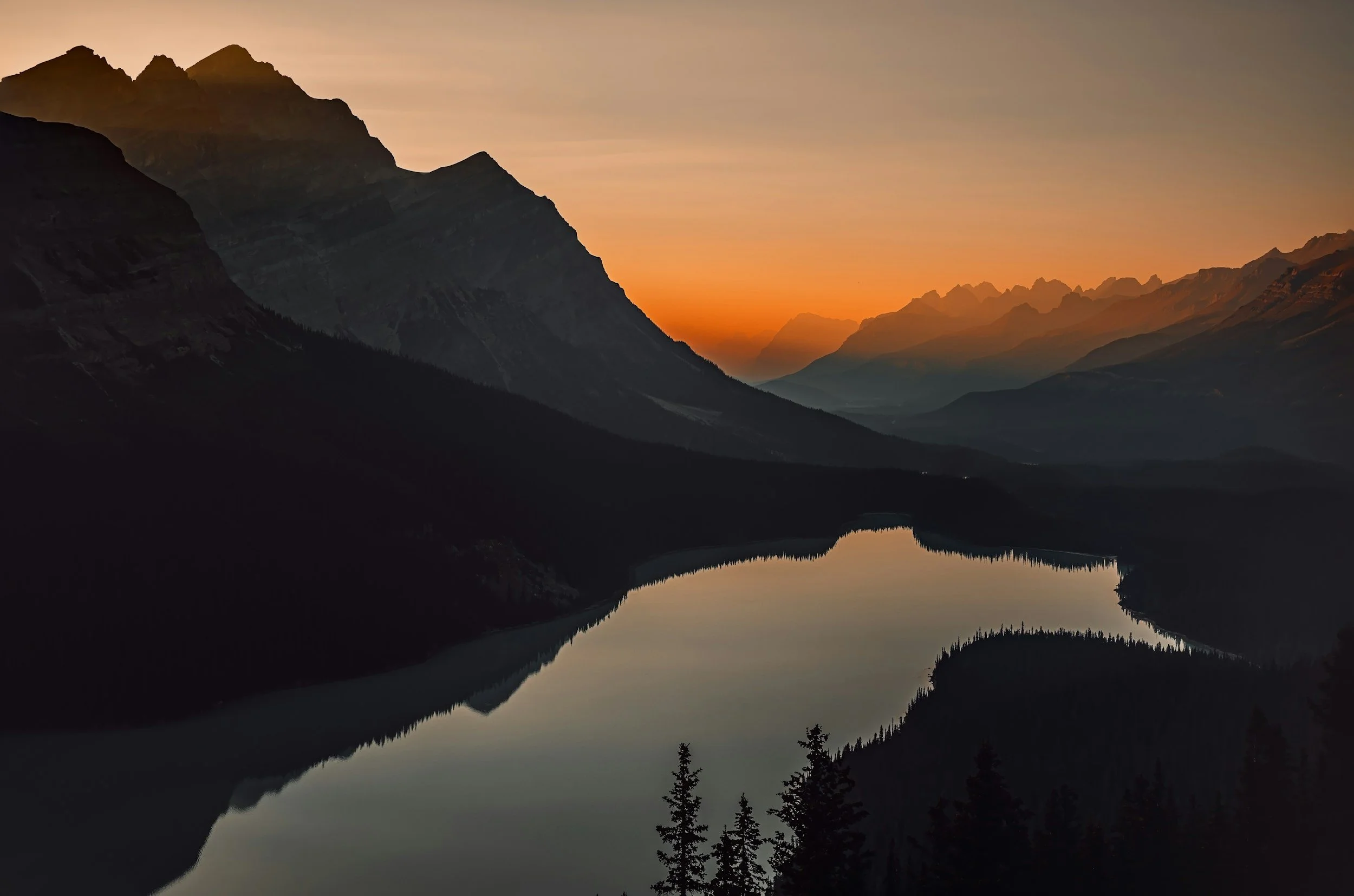A tranquil mountain landscape at sunset with a calm lake reflecting the surrounding mountains and a colorful orange sky.