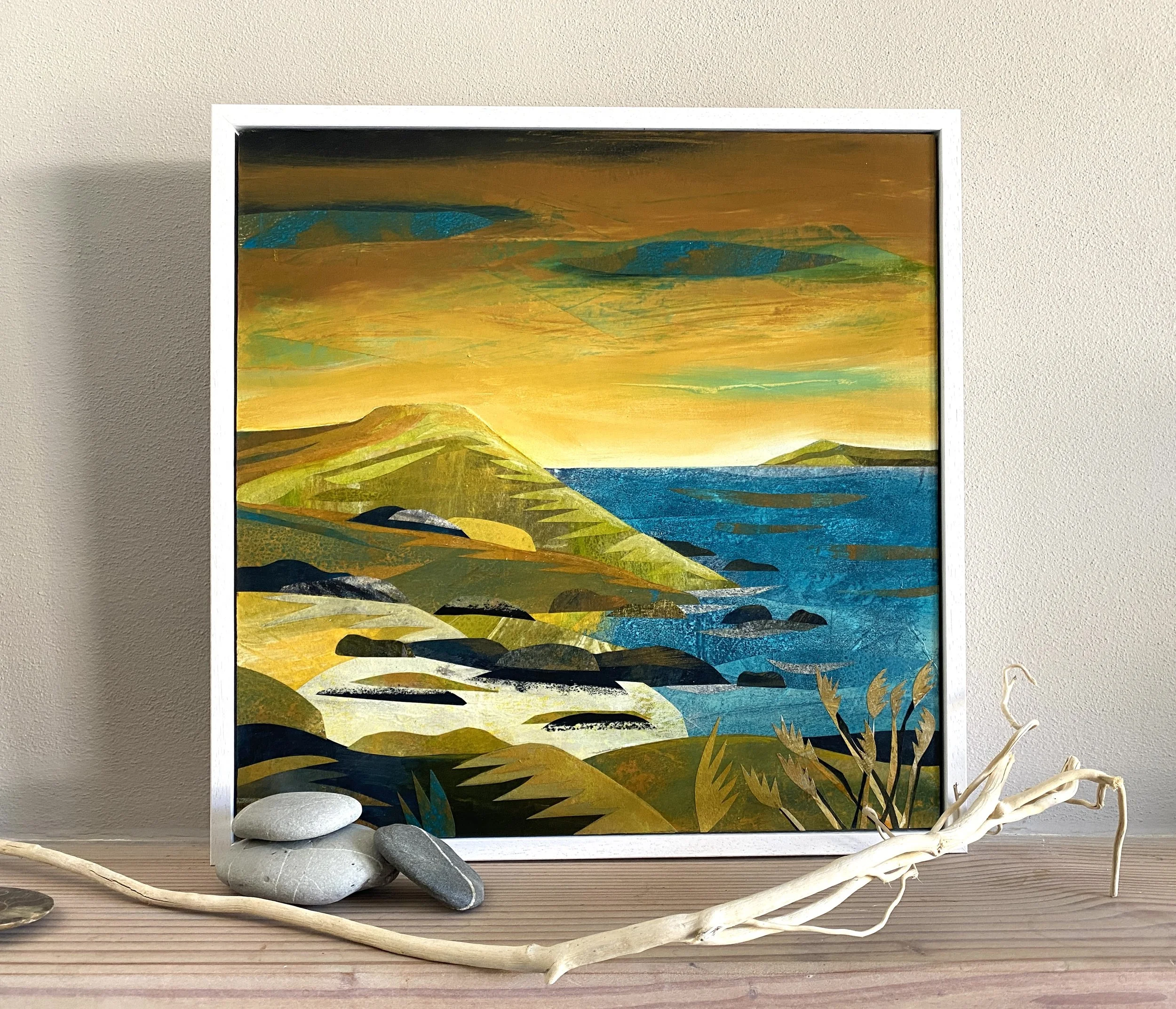 A framed landscape painting of a coastal scene with green hills, rocks, blue water, and a sunset sky. Decorative stones and a white driftwood branch are placed in front of the frame.