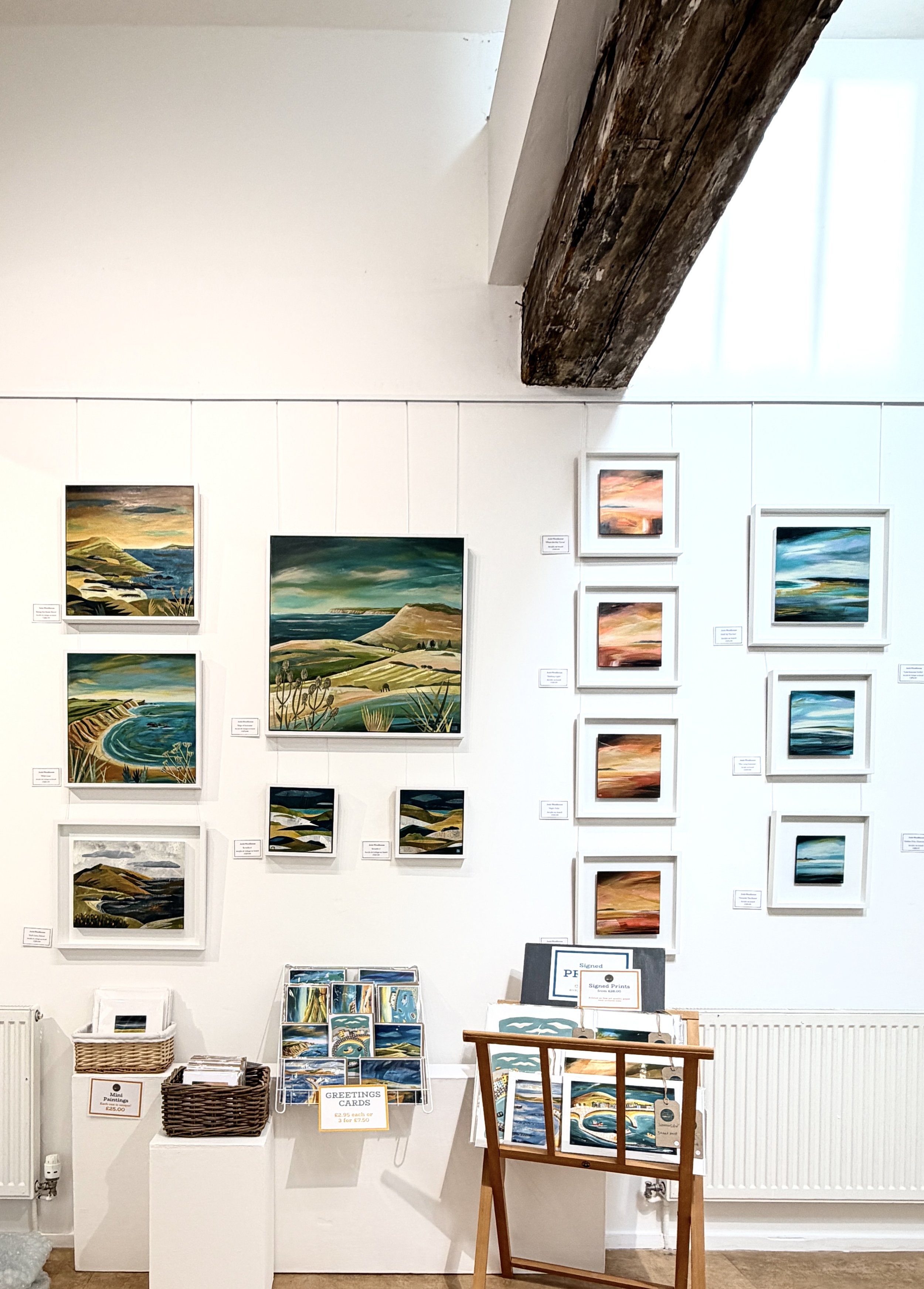Gallery wall featuring landscape paintings by Jools Woodhouse of coastal and hillside scenery, arranged with small and large framed art. Beside the artwork, there is a table with greeting cards and prints for sale.
