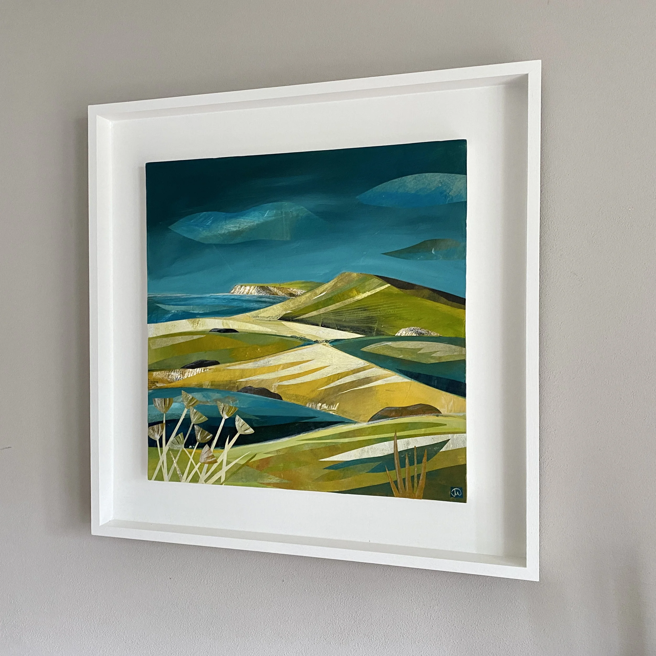 Colorful landscape painting of rolling hills, fields, and a cloudy sky in a white frame hanging on a gray wall.