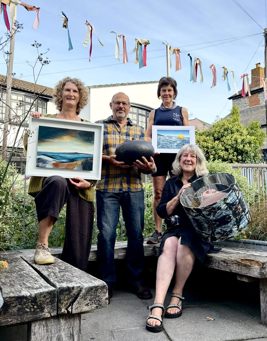 Group of five people outdoors, holding framed landscape paintings and decorative objects, with colorful fabric banners hanging overhead and a garden setting in the background.