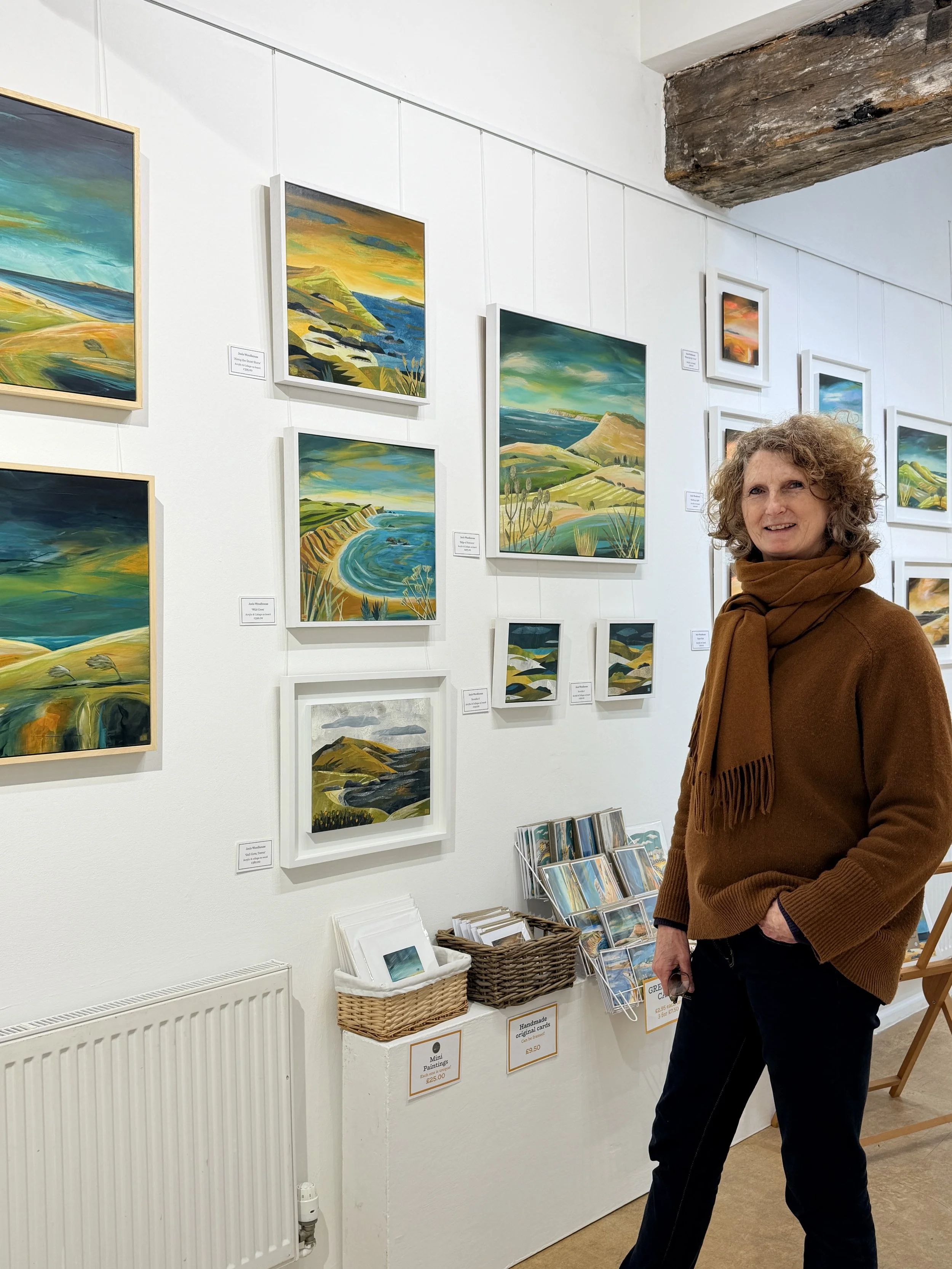 Artist with curly hair wearing a brown sweater and scarf, standing in an art gallery with landscape paintings on white walls.