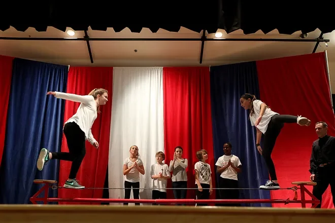 A man in a blue t-shirt helping a boy in a white t-shirt balance on a tightrope during a summer camp activity, with other children in the background participating in similar activities.