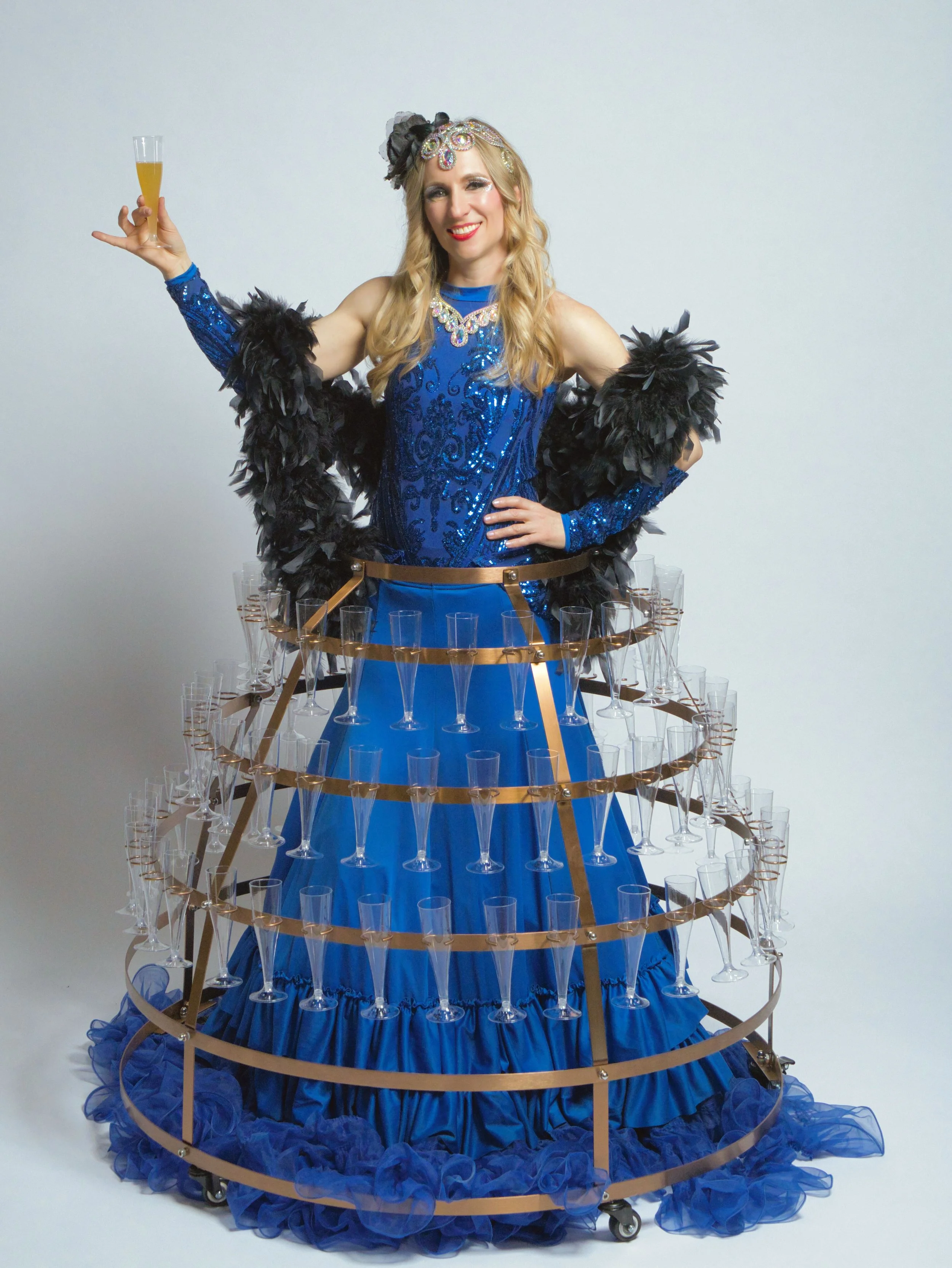 A woman in a blue ball gown, decorated with sequins, stands inside a golden wheel of champagne glasses, holding a glass of champagne, and smiling at the camera.