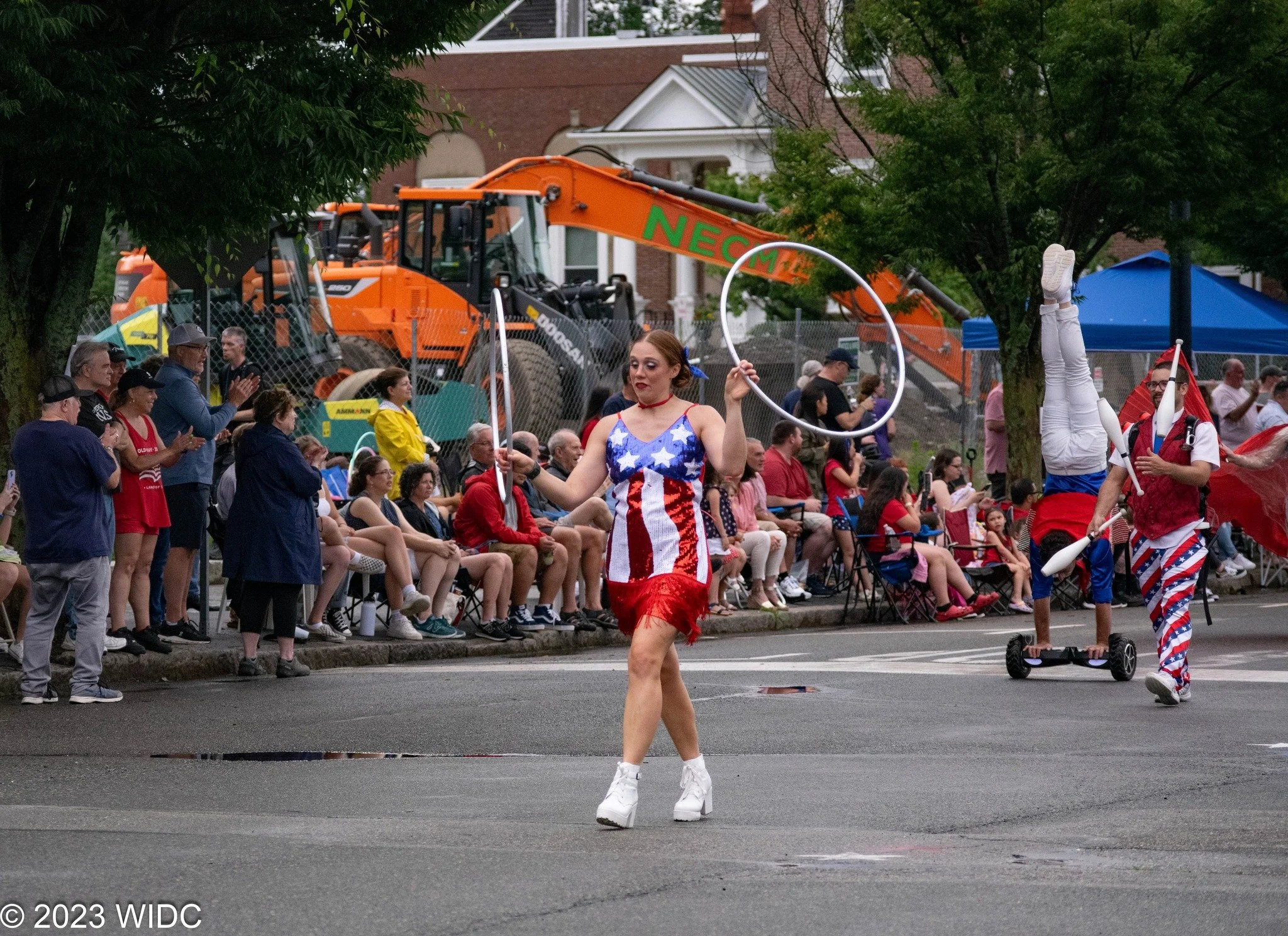 A parade featuring performers dressed in patriotic outfits, with an audience seated along the sidewalk. One woman is spinning a hoop, wearing a dress with the American flag design, and other performers are balancing unicycles and spinning pins. Construction equipment is visible in the background.