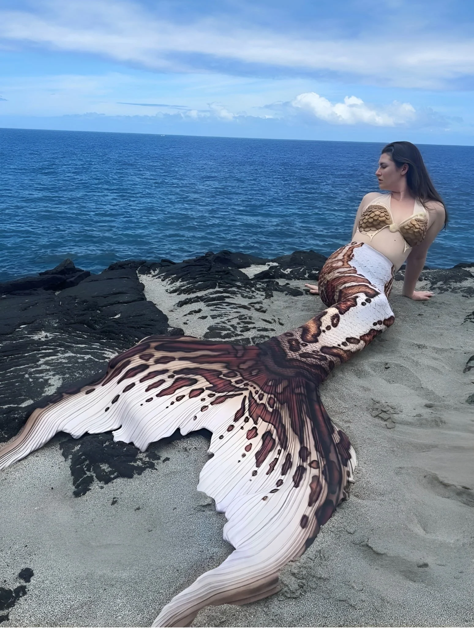 A woman dressed as a mermaid sitting on a rock by the ocean, with her tail extended on the sand.