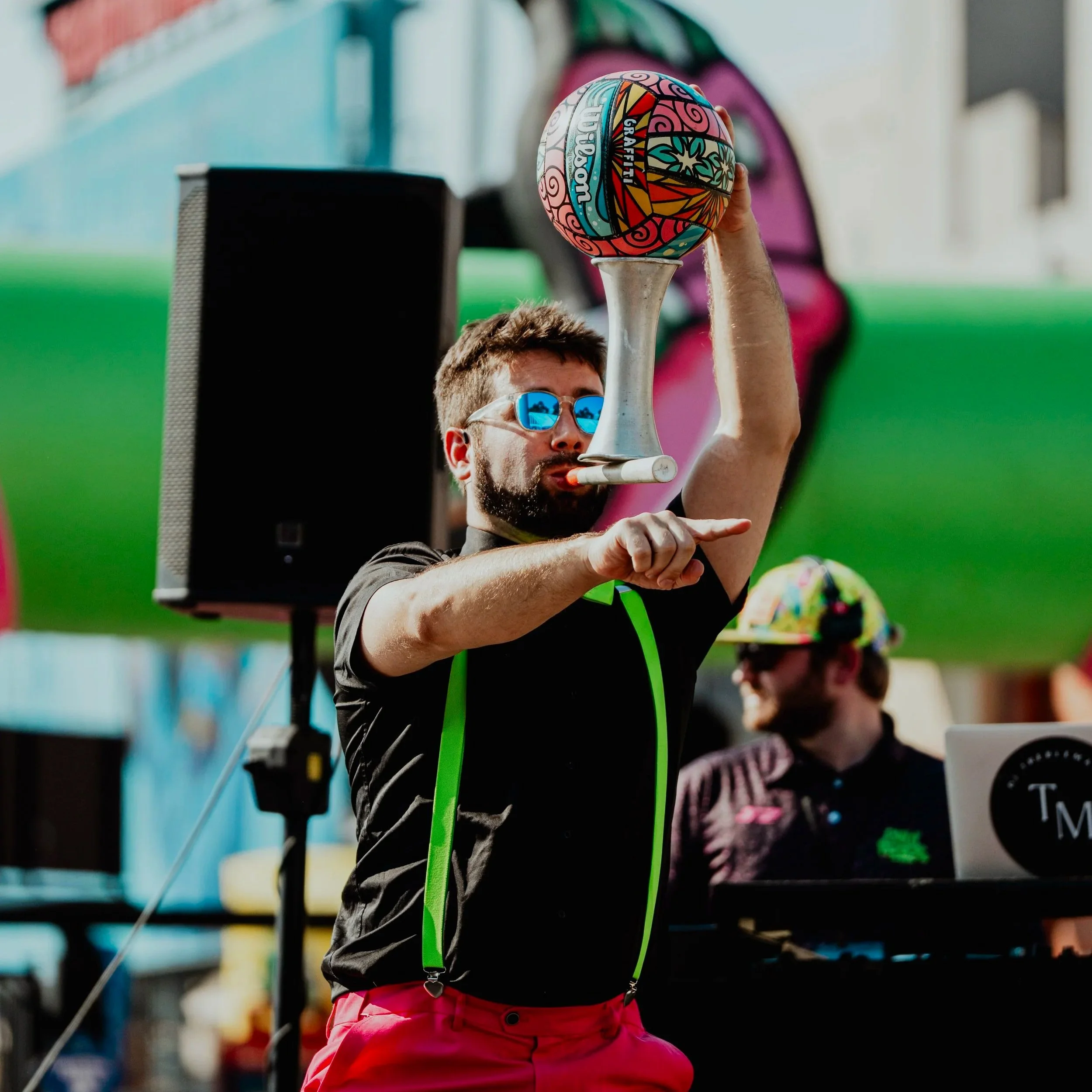A man wearing sunglasses, a black shirt with green suspenders, and red pants is performing a diabolo trick with a colorful ball in an outdoor setting. Another person with a hat is in the background near a DJ setup.
