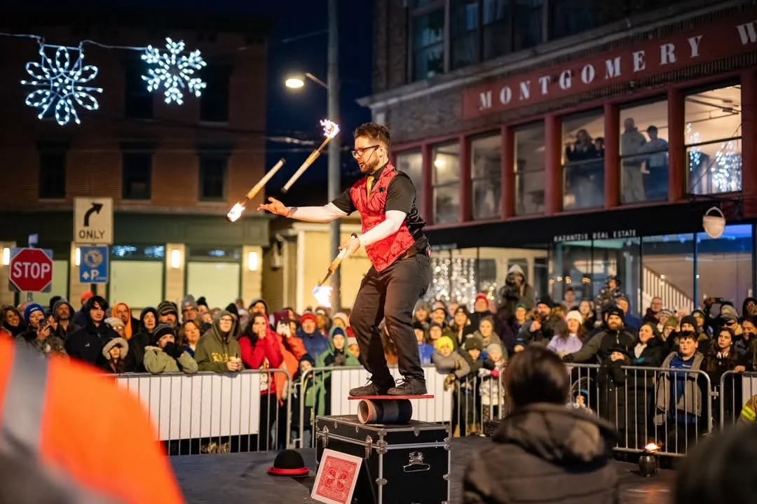 A street performer juggles flaming torches on a large rolling hoop in a busy outdoor entertainment event at night. A crowd of spectators watches behind metal barriers, bundled in warm clothes, on a city street decorated with holiday lights and snowflake ornaments.