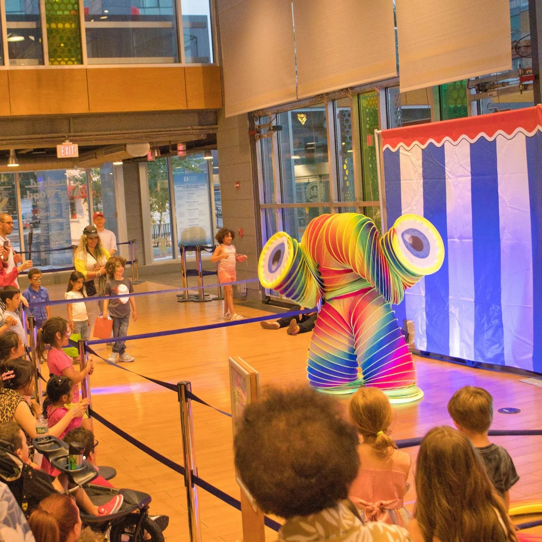 A colorful clown performes in front of an audience of children, with rainbow-colored layers and large circular eye pieces, in an indoor entertainment venue with wooden floors and large windows.