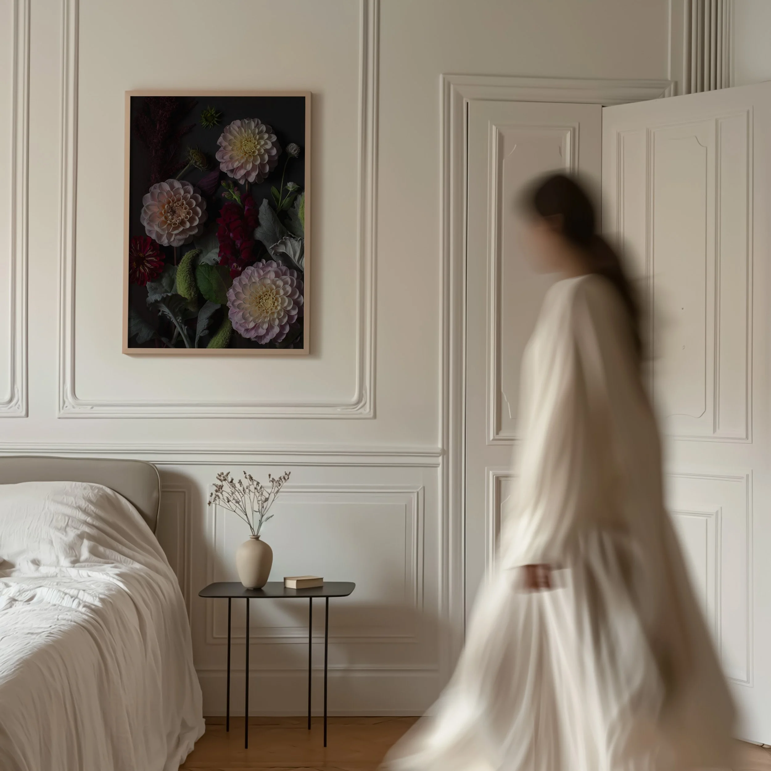 A blurred woman walking past a bedroom with white walls, a bed with white bedding, a black side table, a white vase with dried flowers, and a framed floral art piece on the wall.