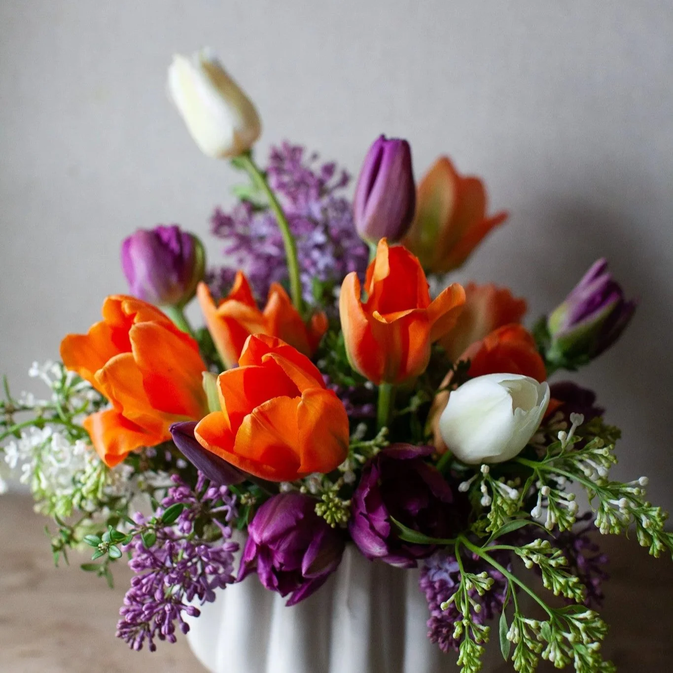 Close-up of a colorful bouquet of various flowers in a glass vase, including pink tulips, yellow tulips, white anemones, and other mixed flowers with greenery.