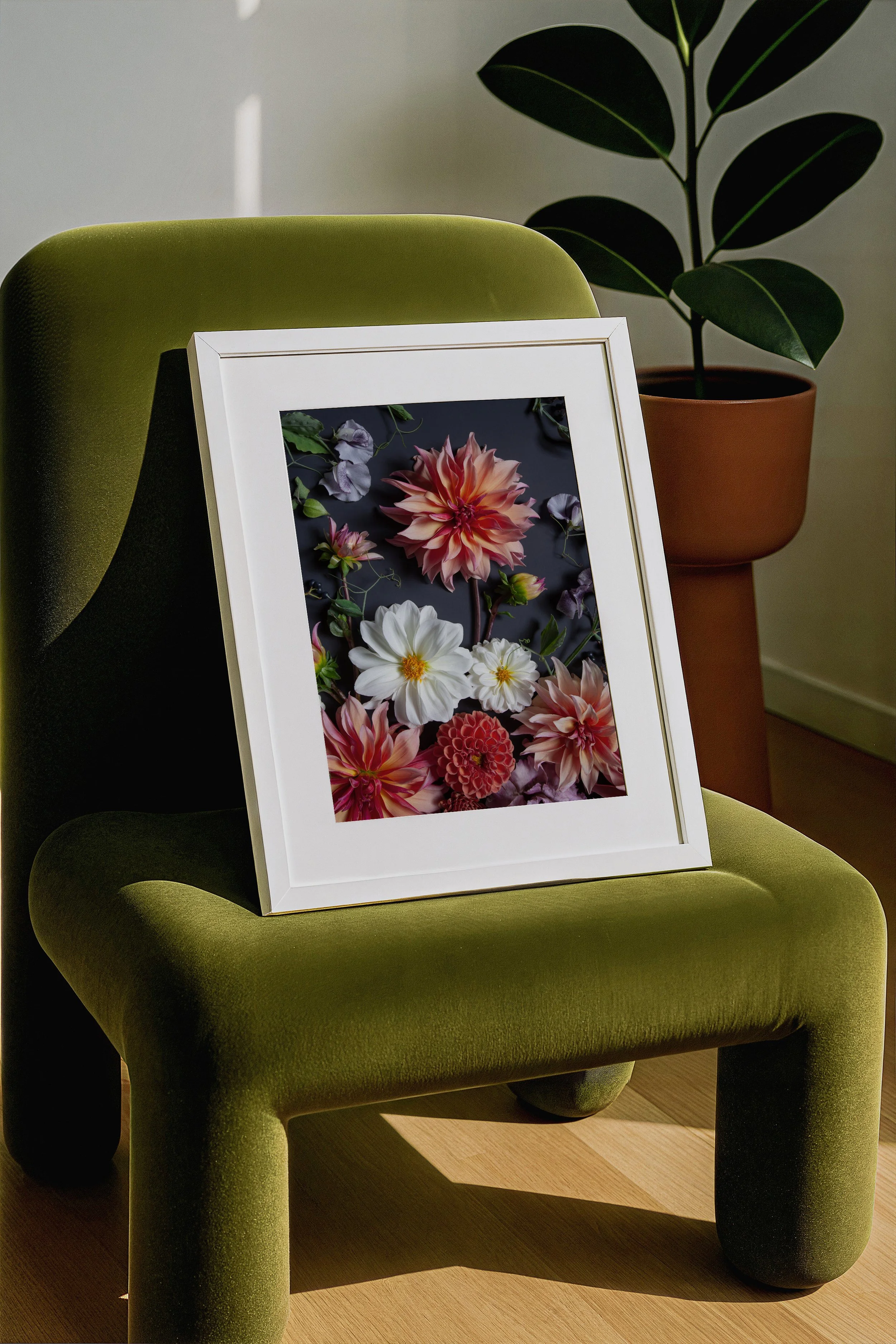 A white picture frame with a floral photo resting on a plush green velvet chair, with a potted plant in the background.