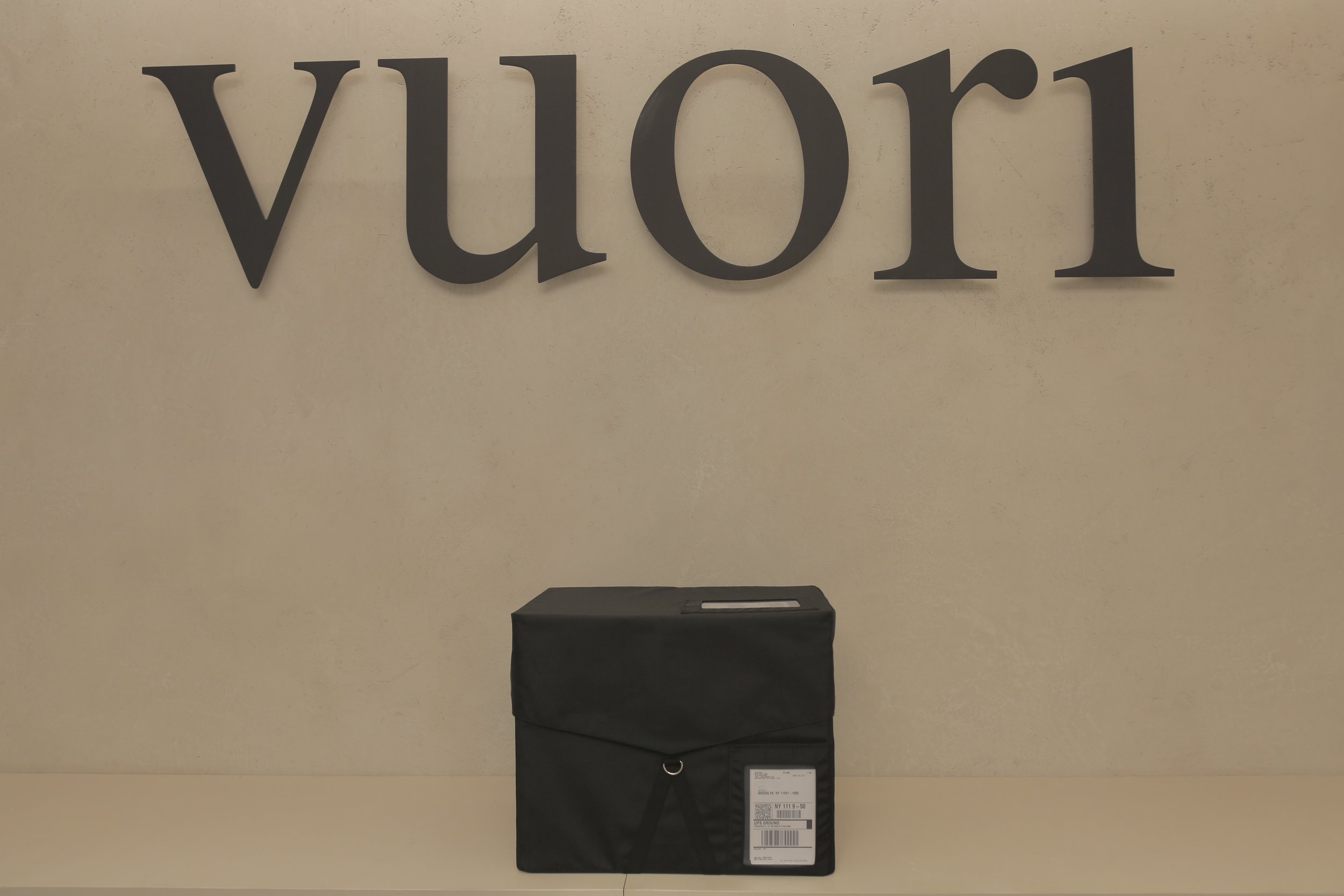 A black courier bag with a handle and a shipping label, placed on a beige table against a wall with the large black word 'VUIOR' on it.