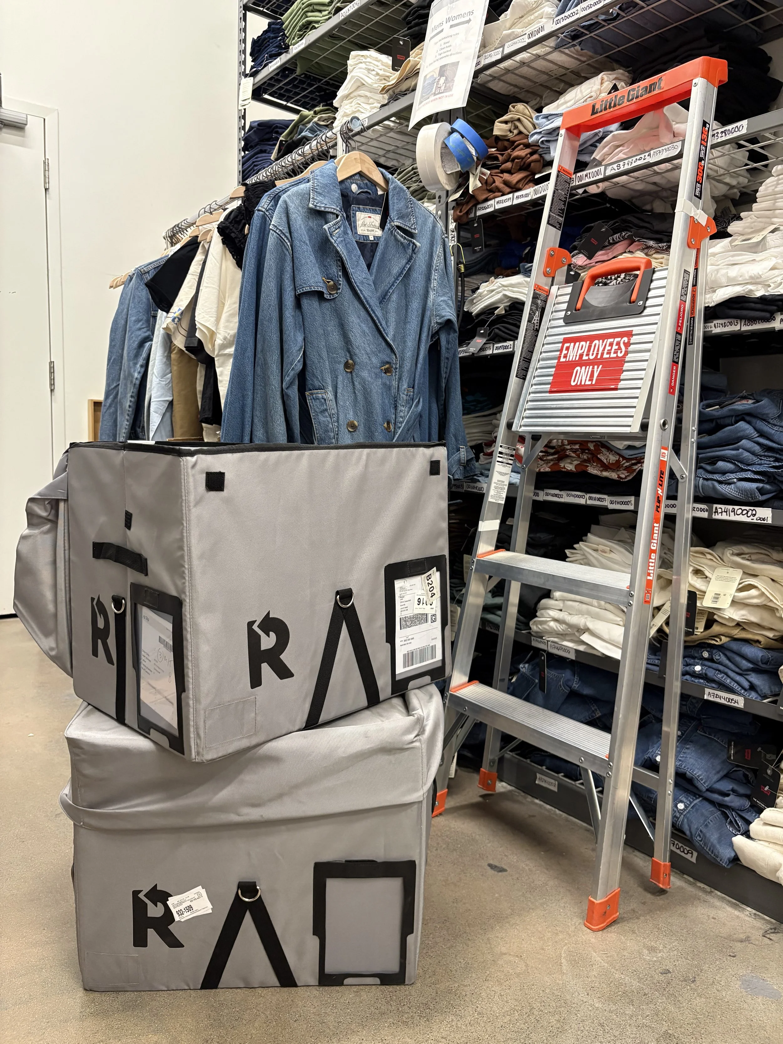 Clothing store display with shelves of folded clothes, a denim jacket hanging on a rack, and two large gray boxes with black labels and handles, a small step ladder with a red "Employees Only" sign in front of the shelves.