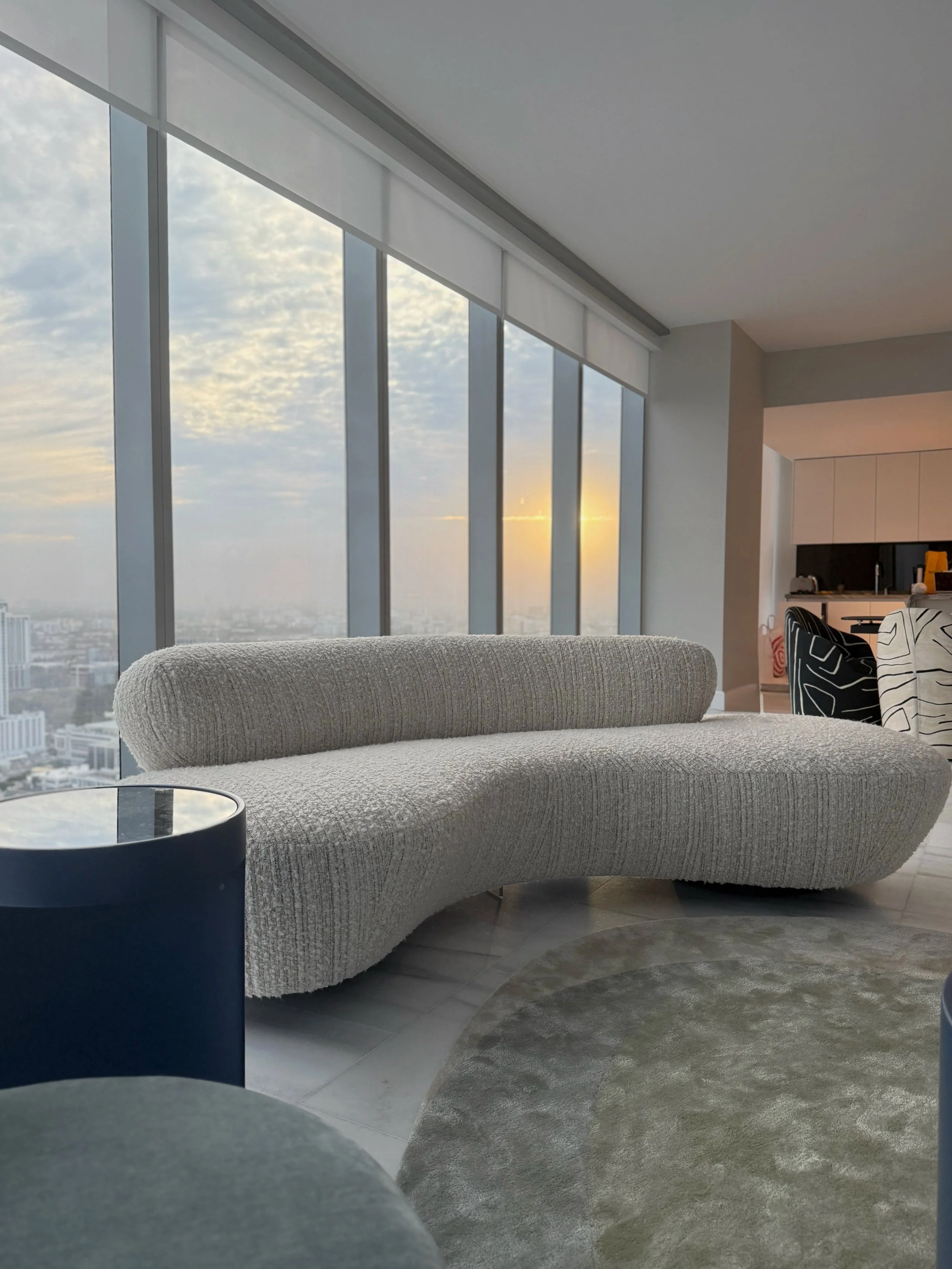 A modern penthouse living room, such as in Fisher Island, Miami reflects refined coastal luxury with expansive floor-to-ceiling windows framing sweeping water views. Curved contemporary sofas, plush textured fabrics, and sculptural lighting establish