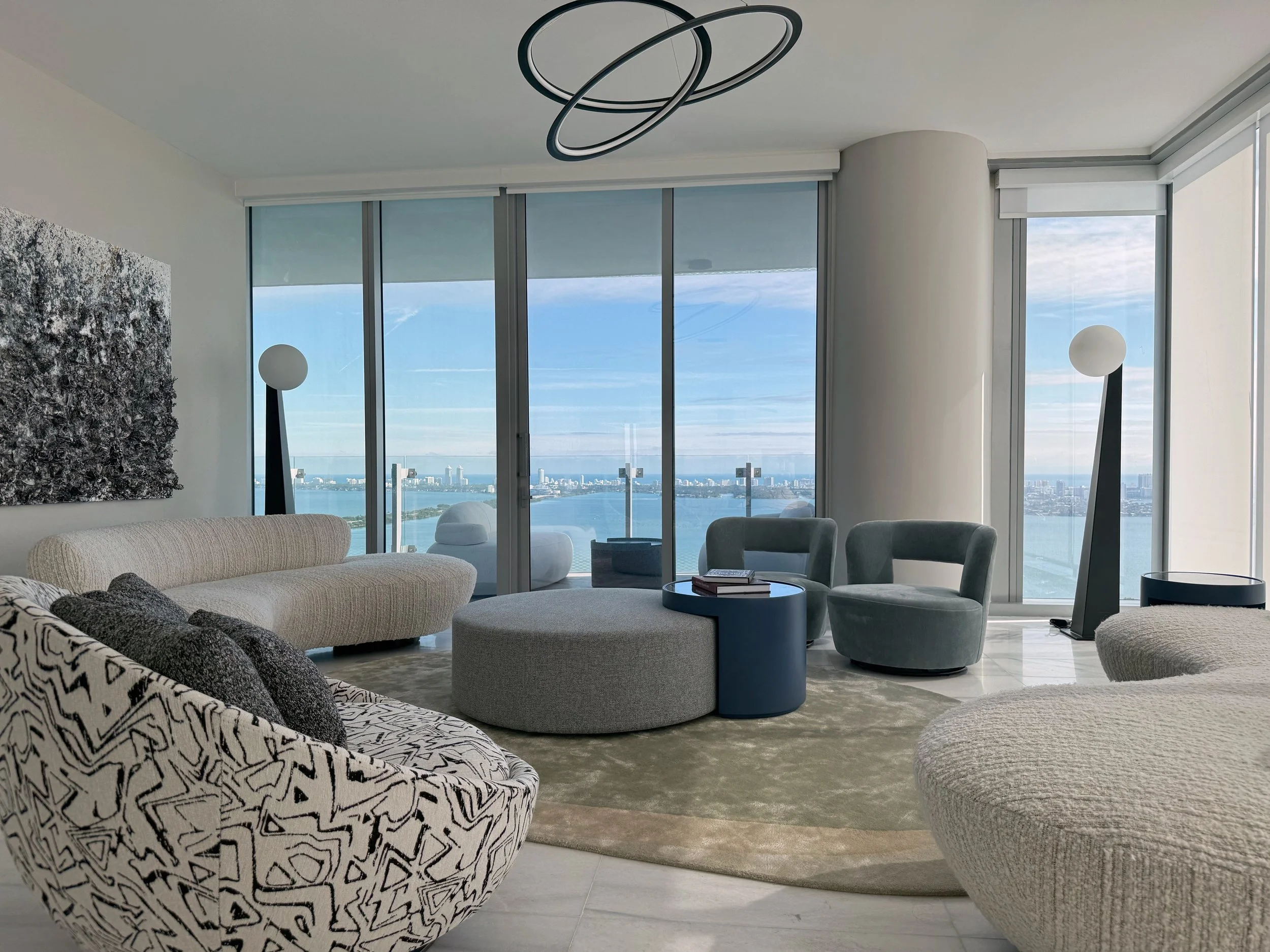 An elegant penthouse interior, such as in Coconut Grove, Miami showcases modern luxury condo design centered around large glass windows that frame spectacular waterfront views. A composition of curved modern seating, plush textured upholstery, and mi