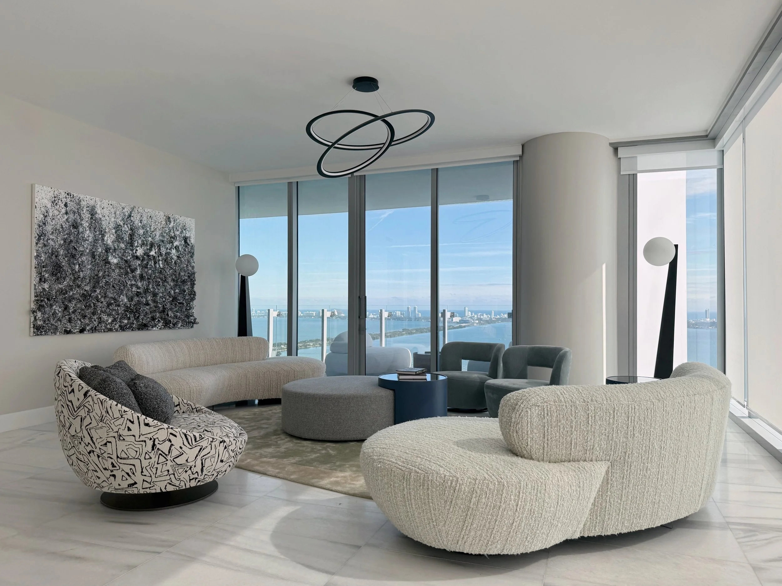 Modern penthouse living room in Edgewater, Miami showcasing contemporary luxury interior design with sweeping floor-to-ceiling windows overlooking Biscayne Bay and the Miami skyline. Sculptural curved sofas, boucle upholstery, and minimalist lighting