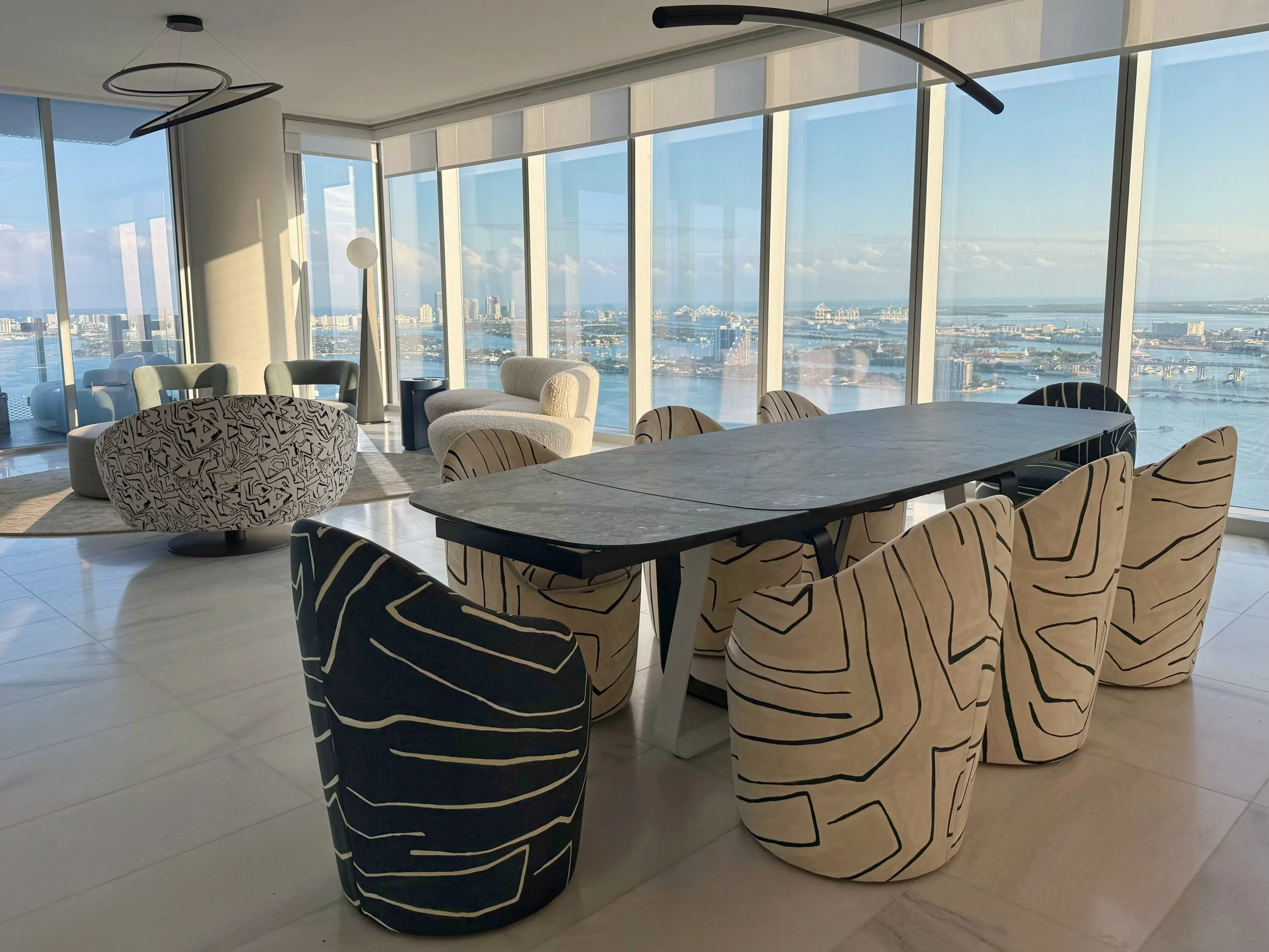 A luxury penthouse dining room in Edgewater, Miami showcasing modern interior design tailored for high-rise condo living with dramatic floor-to-ceiling windows framing sweeping Biscayne Bay and Miami skyline views. A sleek contemporary marble dining 