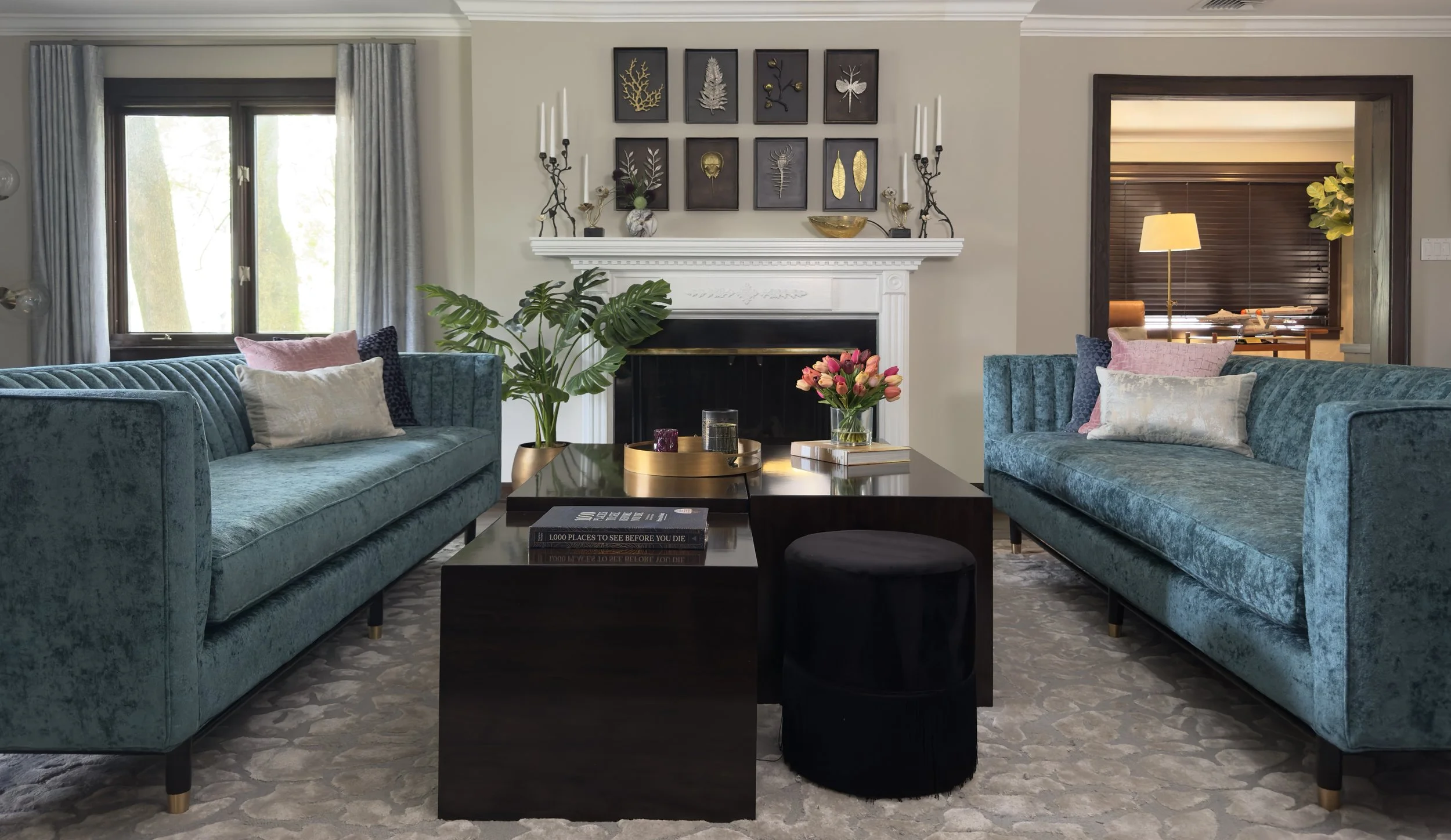 Living room with two blue velvet sofas, a black coffee table decorated with books and flowers, a white fireplace with framed botanical art above, and a window with light blue curtains.