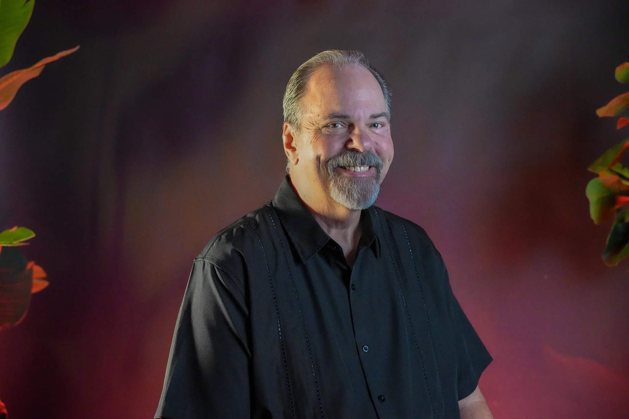 Portrait of a man with short dark hair, a beard and mustache, smiling, wearing a dark blazer and light blue collared shirt, against a beige floral patterned background.
