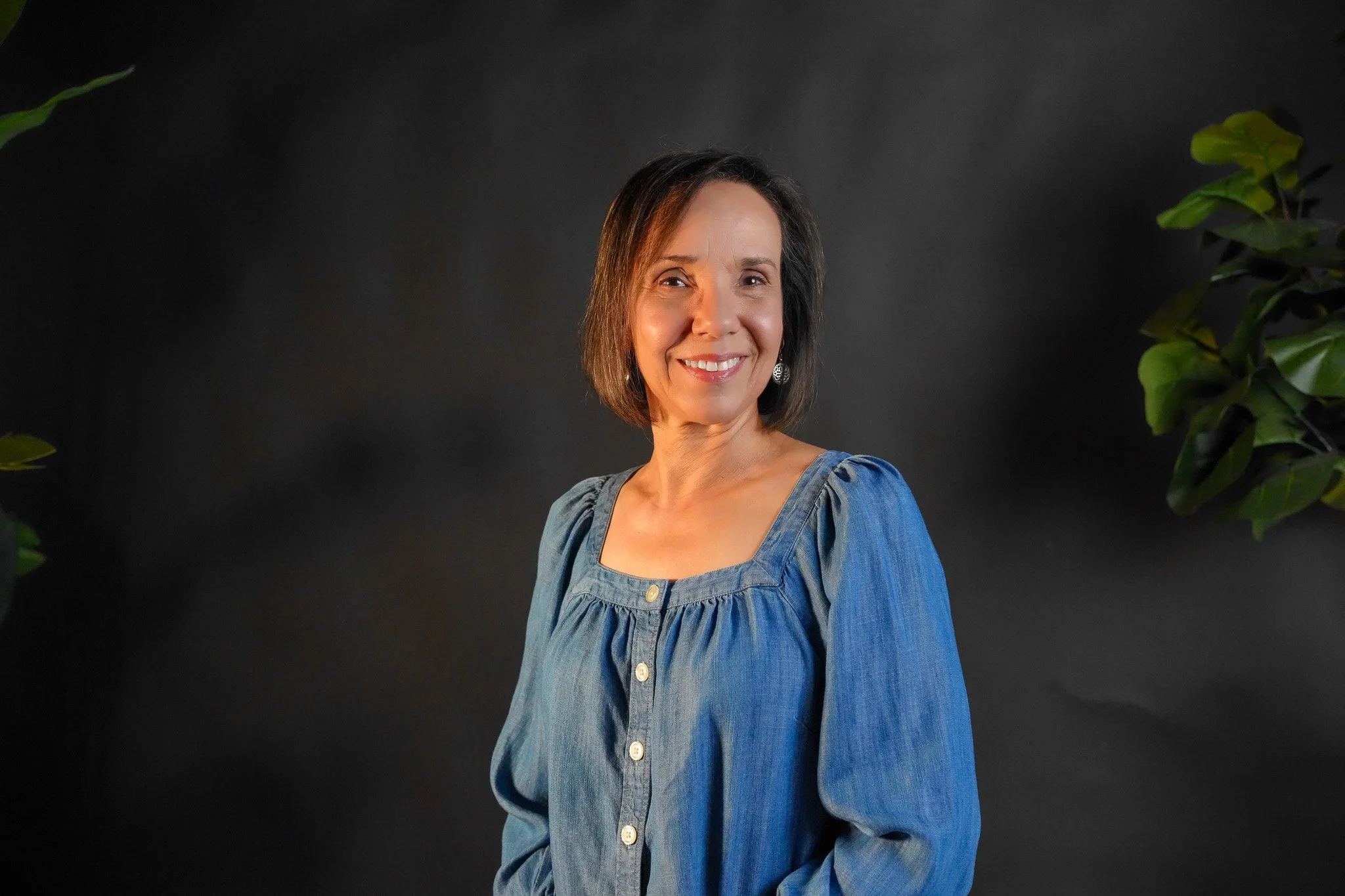 A woman with short brown hair wearing silver earrings and a black blouse, smiling against a black background.