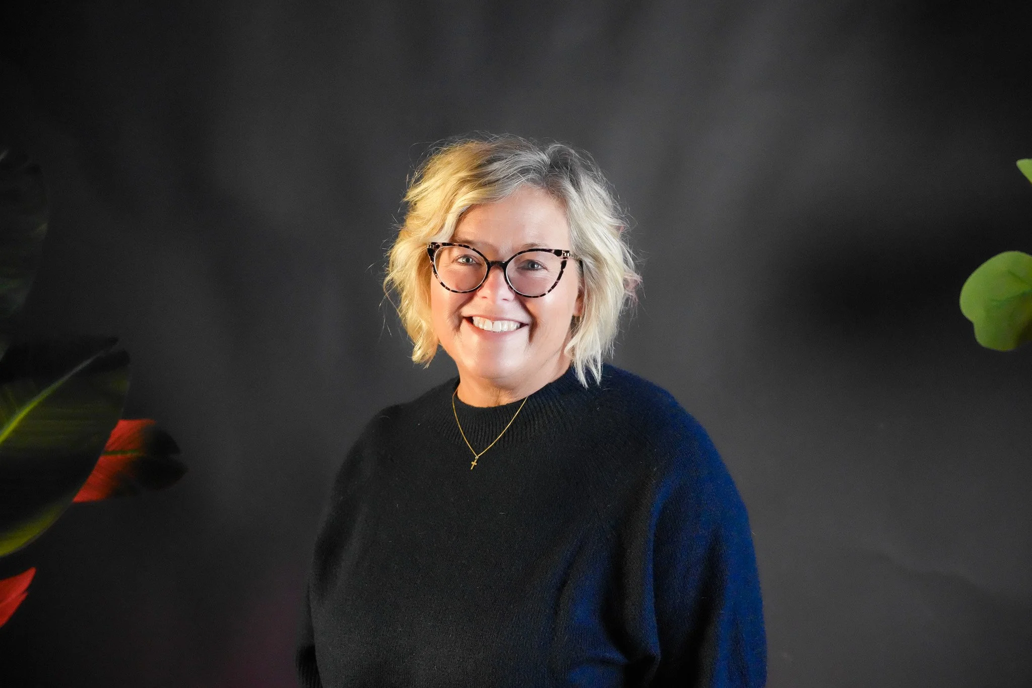 Close-up of a smiling woman with blonde hair, wearing white glasses, a black shirt, a gold necklace with a cross pendant, and wearing earrings. There is colorful floral artwork in the background.