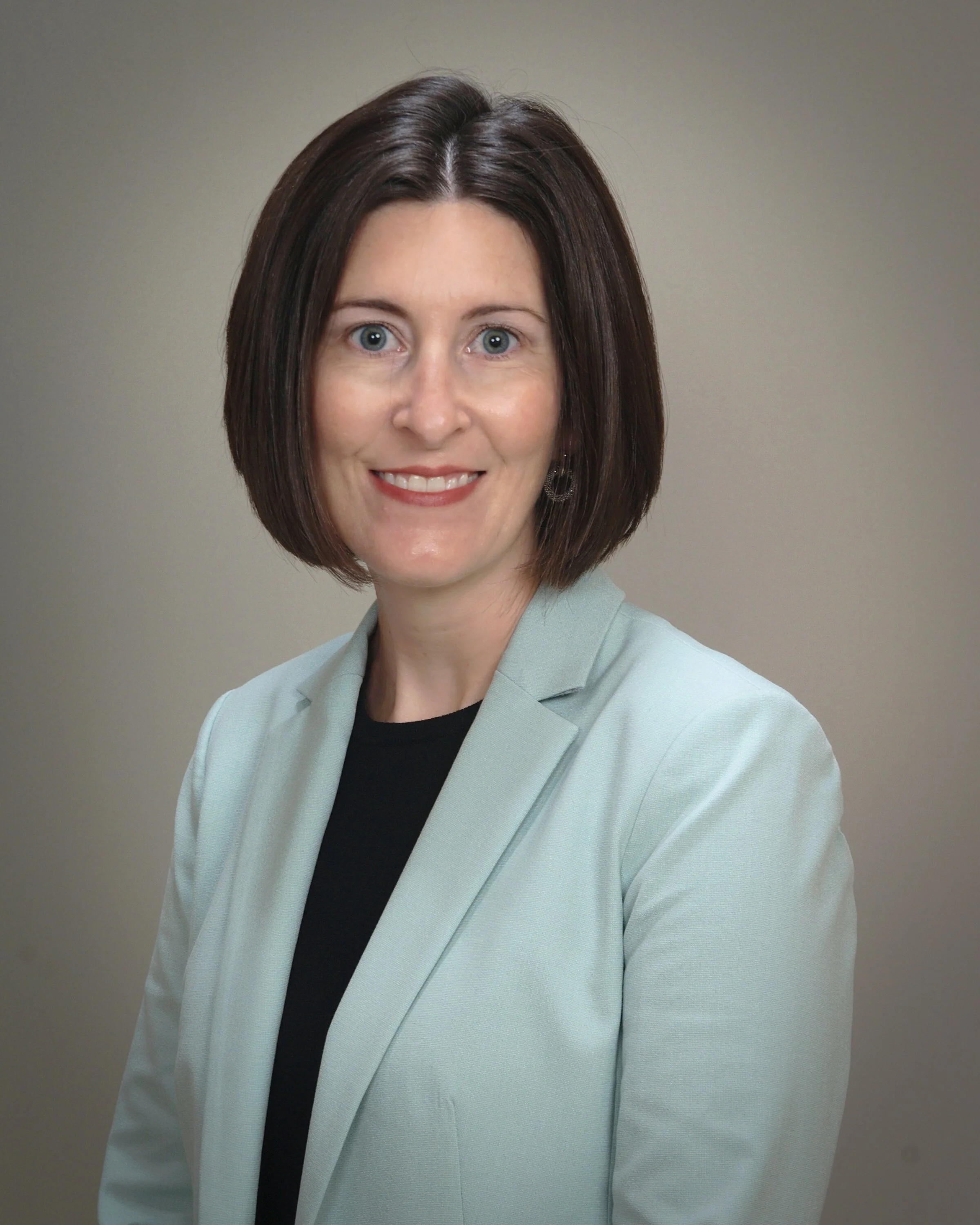Portrait of a woman with shoulder-length dark brown hair, wearing a light-colored blazer over a black top, smiling against a plain gray background.